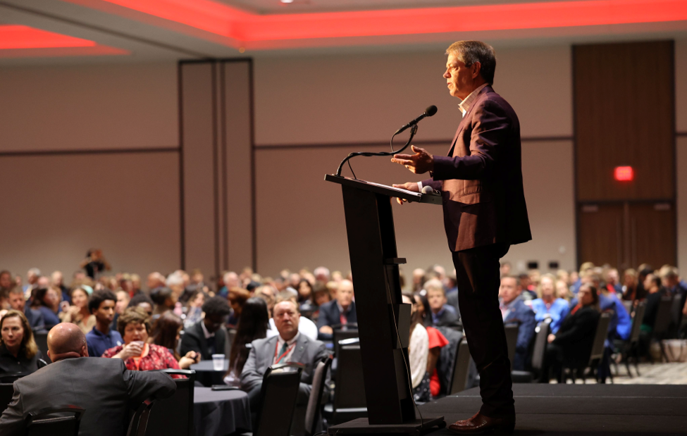 Gov. Pillen delivers opening remarks at Governor’s Summit in Kearney to a room full of people sitting at circular tables with black tablecloths.