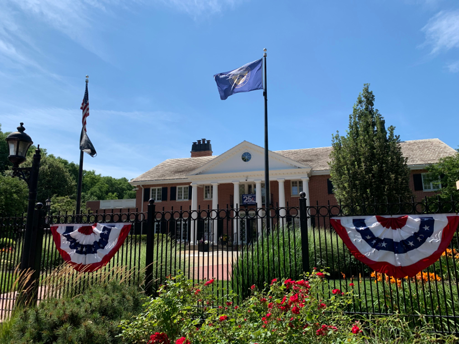 Image of the Governor's Mansion. The fence is adorned with red, white, and blue decorations and flowers. A Nebraska flag flies between the fence and mansion.