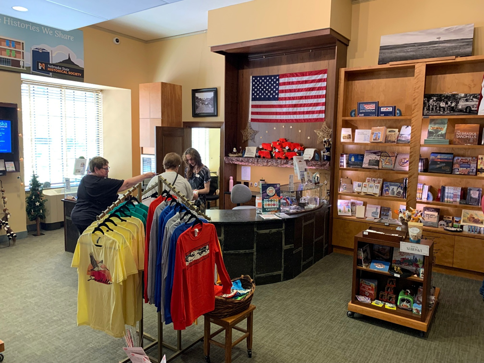The State Capitol Gift Shop with various items for sale, including tee-shirts, books, and candy. Two visitors are at the checkout counter being helped by a gift shop employee.