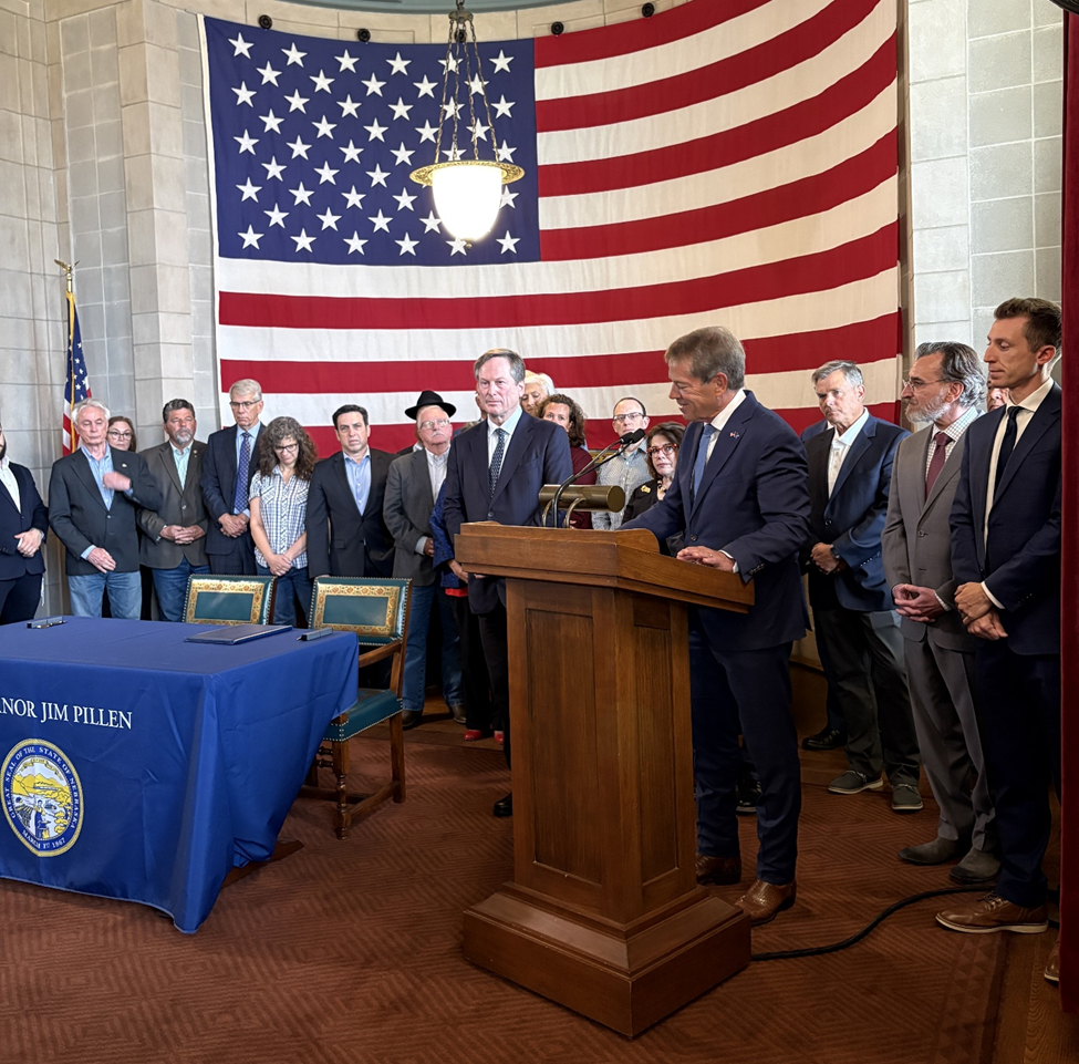 Gov. Pillen speaks from a podium beside a table with two chairs to the audience while state senators and faith leaders stand behind him in front of a giant American flag.