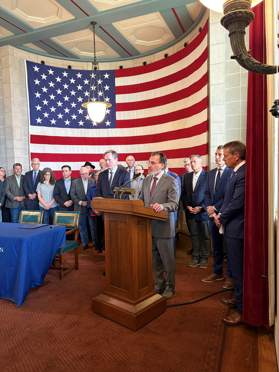 Bob Goldberg from the Jewish Federation of Omaha speaks from a podium beside a table with two chairs to the audience while state senators and faith leaders stand behind him in front of a giant American flag.