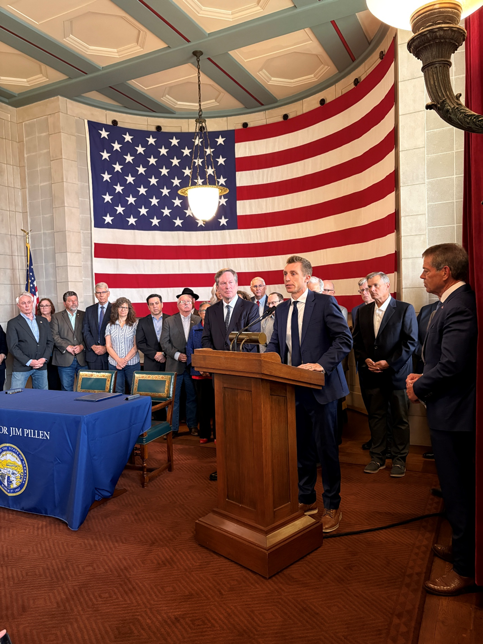 Nate Grasz from Nebraska Family Alliance speaks from a podium beside a table with two chairs to the audience while state senators and faith leaders stand behind him in front of a giant American flag.
