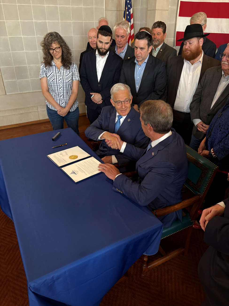 Governor Jim Pillen and Secretary of State Bob Evnen sit at a table congratulating each other after signing the Executive Order in front of state senators and faith leaders.
