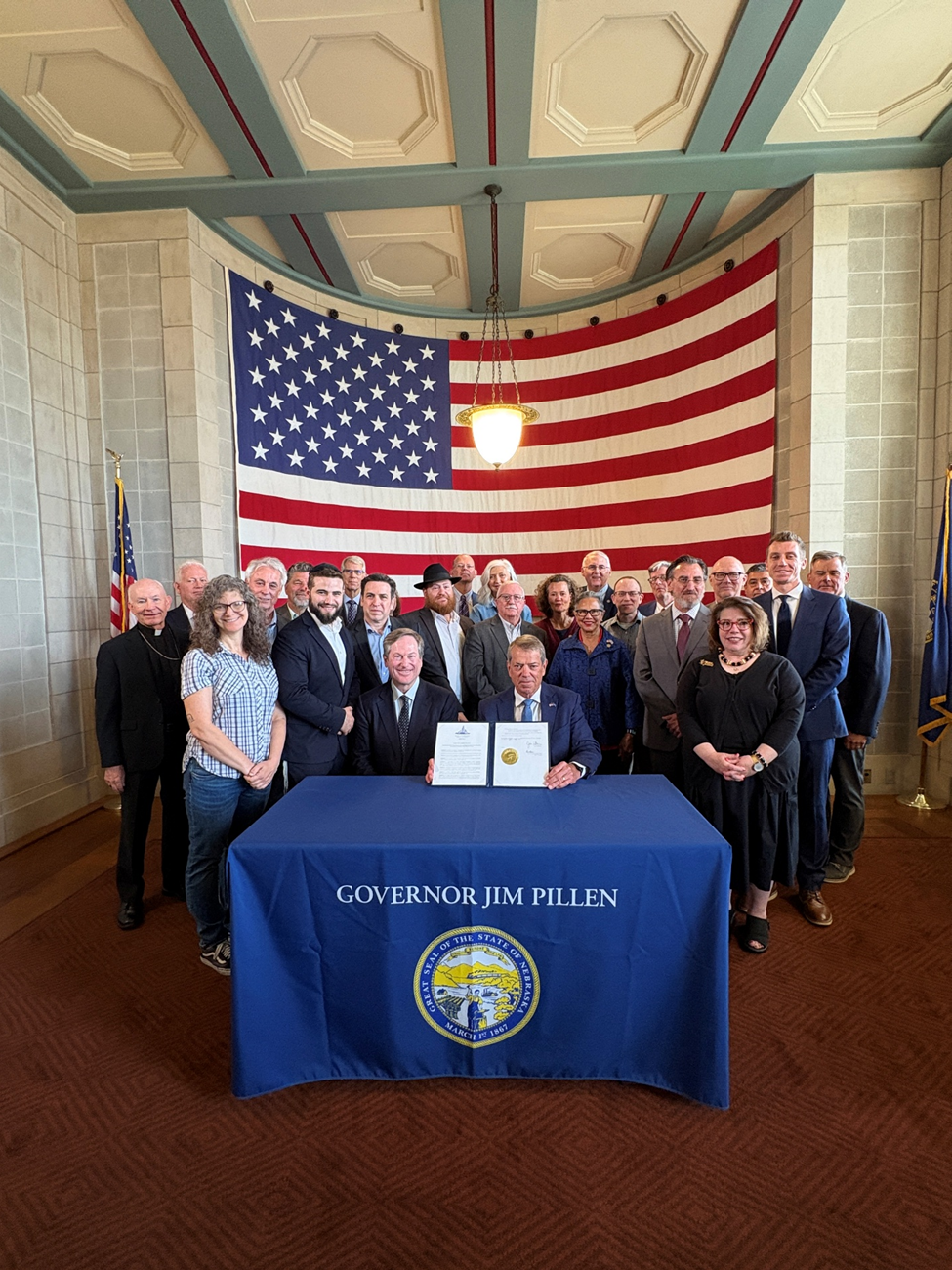 Governor Pillen displays the Executive Order while seated at the signing table and surrounded by state senators and faith leaders with a giant American flag in the background..