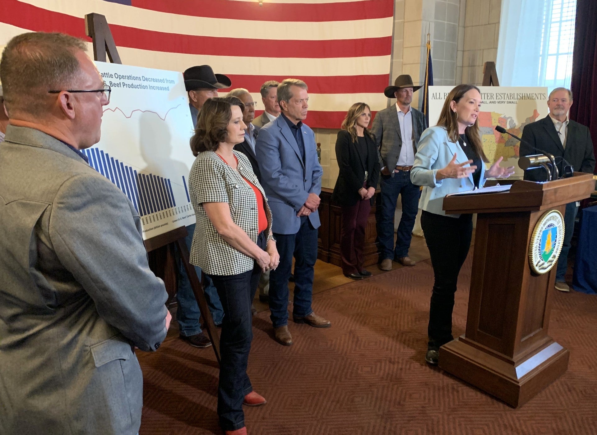 U.S. Department of Agriculture Secretary Brooke Rollins speaks at a podium during a press event, surrounded by cattle industry representatives and officials. An American flag and charts about U.S. beef cattle operations and slaughter establishments are displayed in the background.