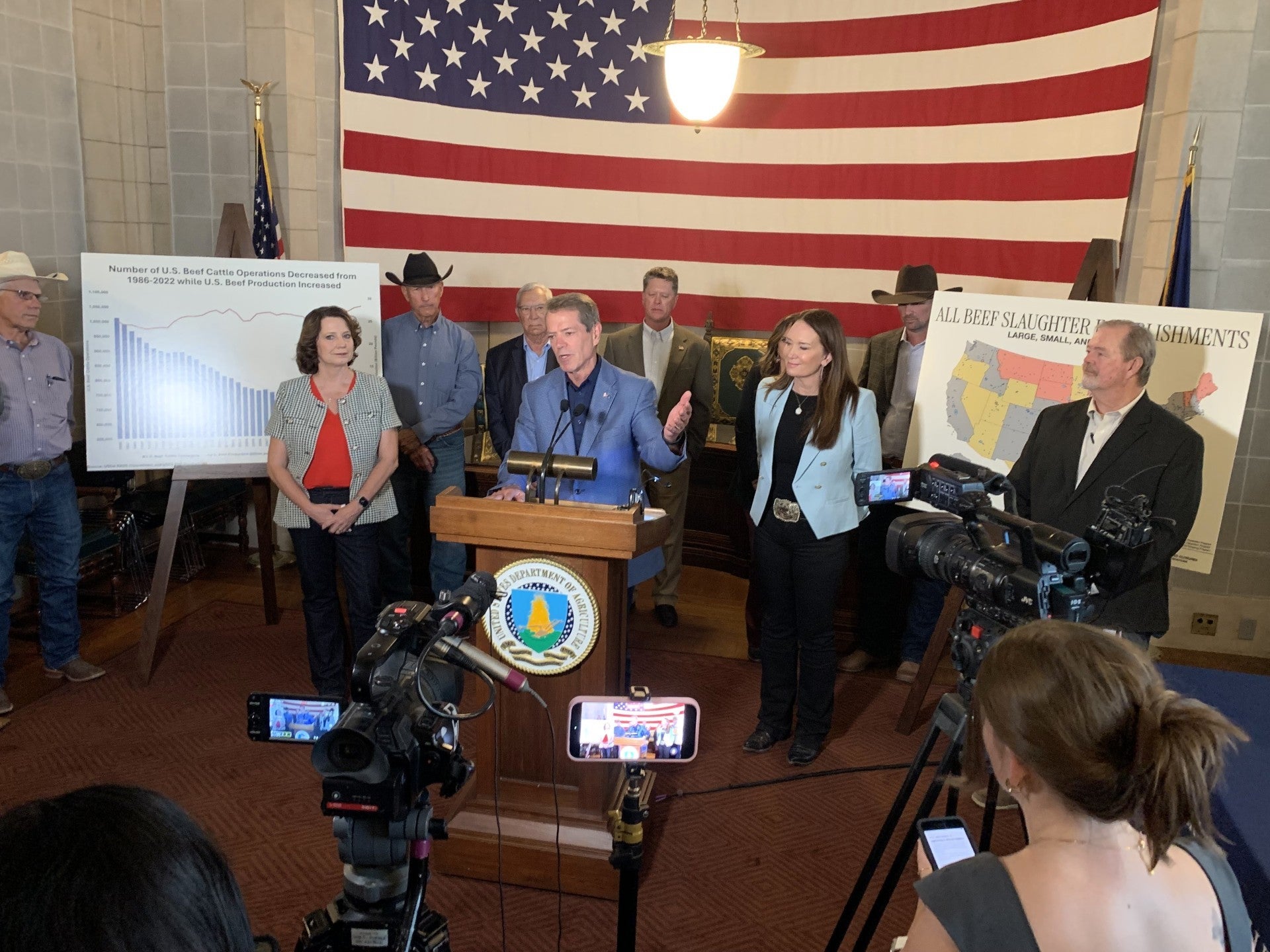 Governor Jim Pillen thanks U.S. Department of Agriculture Secretary Brooke Rollins at a podium during a press conference about the cattle industry, surrounded by officials and industry representatives. News cameras record the event as charts showing declining U.S. beef cattle operations and a map of beef slaughter establishments stand beneath a large American flag.