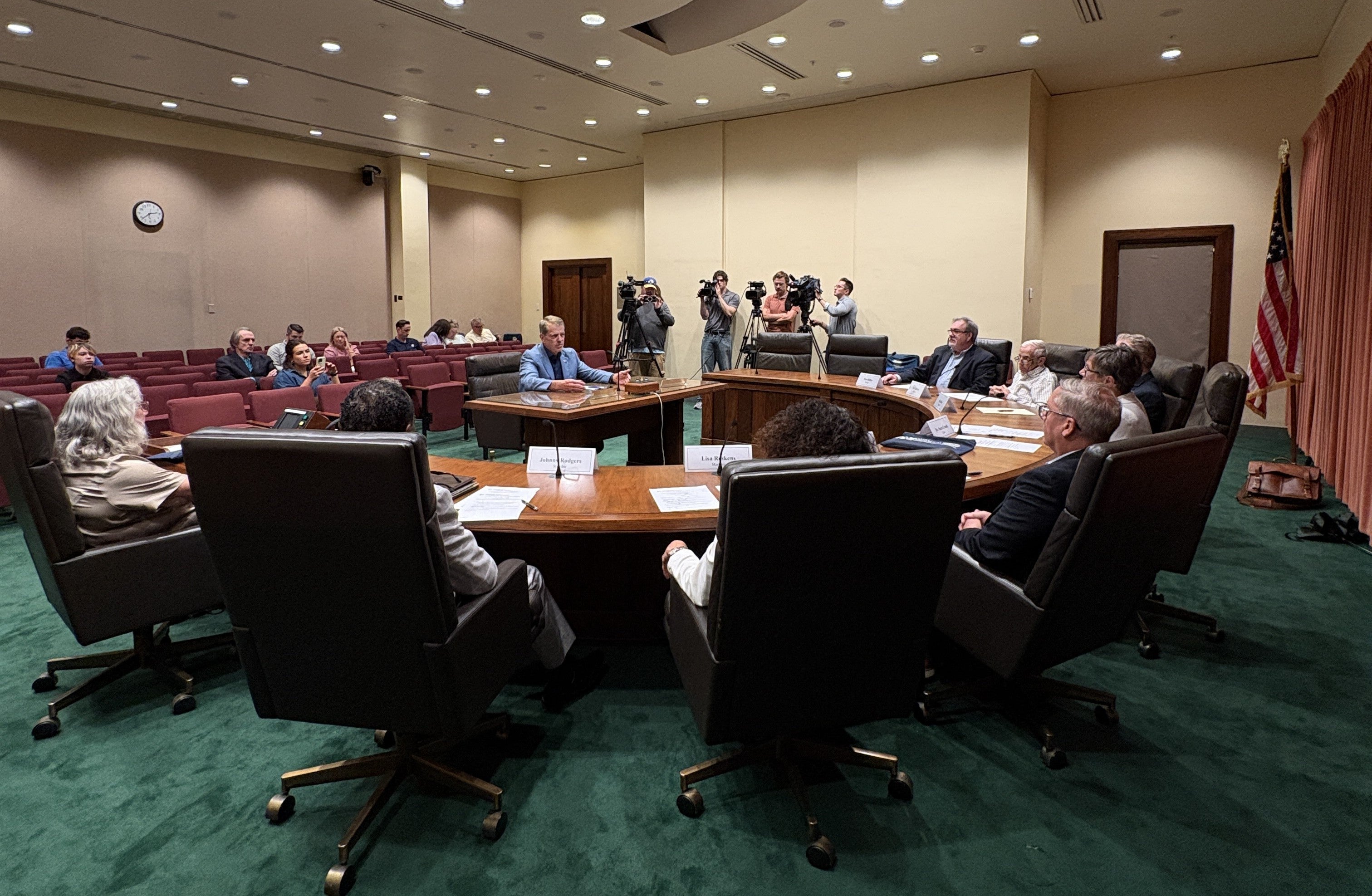 Governor Jim Pillen addressing members of the Nebraska Hall of Fame Commission. Governor Pillen is sitting at a table between members of the commission, who are in seated at a semi-circular table, an audience behind him, and men with cameras to his left.
