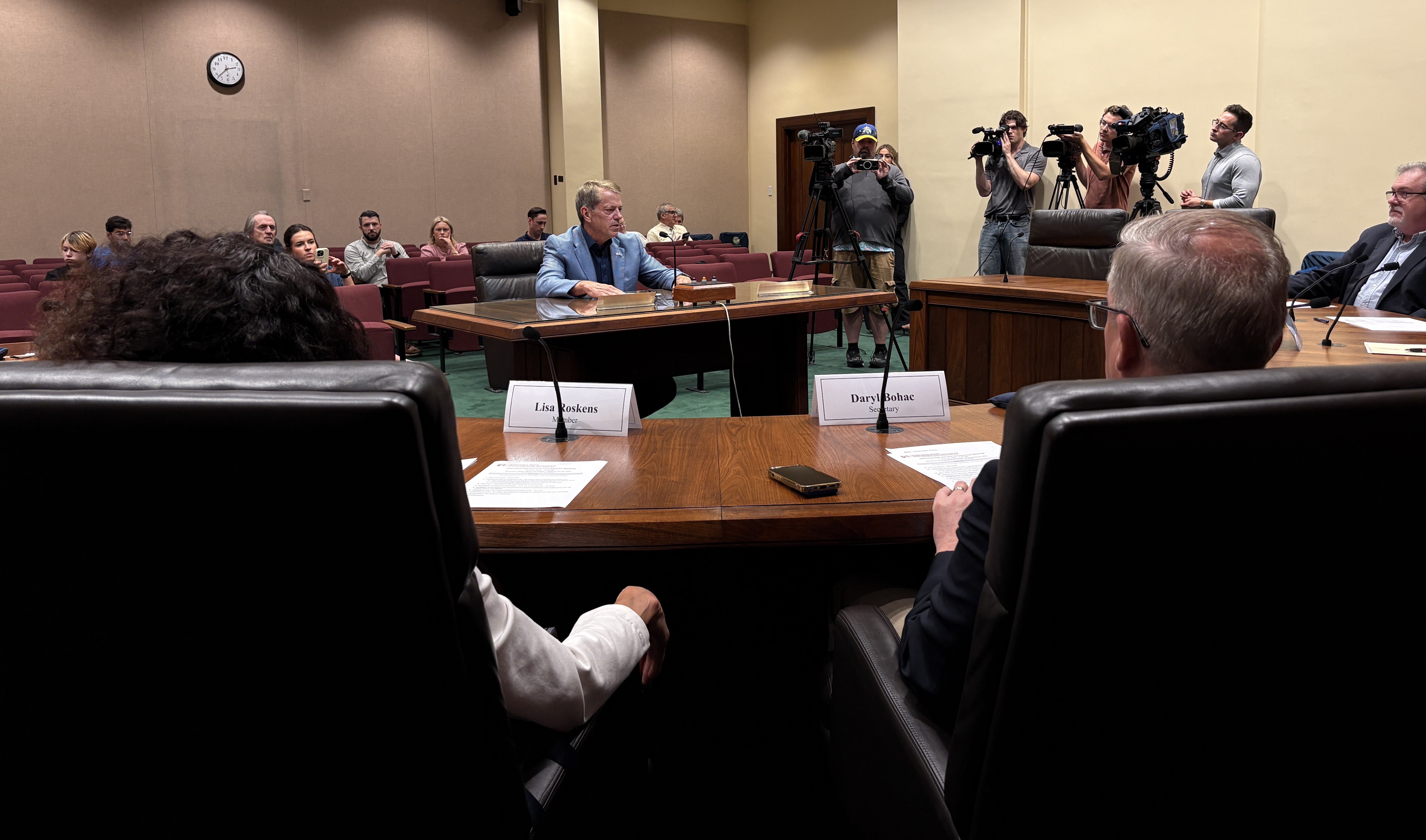 A closer view of Governor Jim Pillen addressing members of the Nebraska Hall of Fame Commission. Governor Pillen is sitting at a table between members of the commission, who are in seated at a semi-circular table, an audience behind him, and men with cameras to his left.