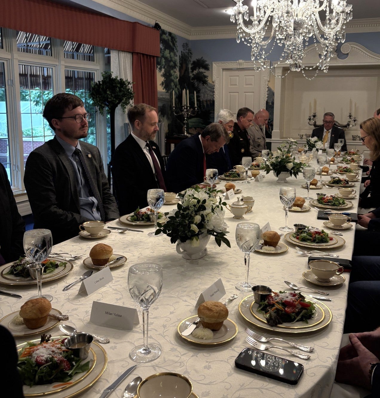 Members of the group join Gov. Jim Pillen and President Petr Pavel in bowing their heads and saying grace around the dinner table in the Governor's Mansion before they eat. There are China plates filled with salad, dinner rolls, and a water glass at every place setting. Dinnerware and tea cups and a name plate adorn each place. Tree pots filled with flowers sit in the middle and each end of the table.