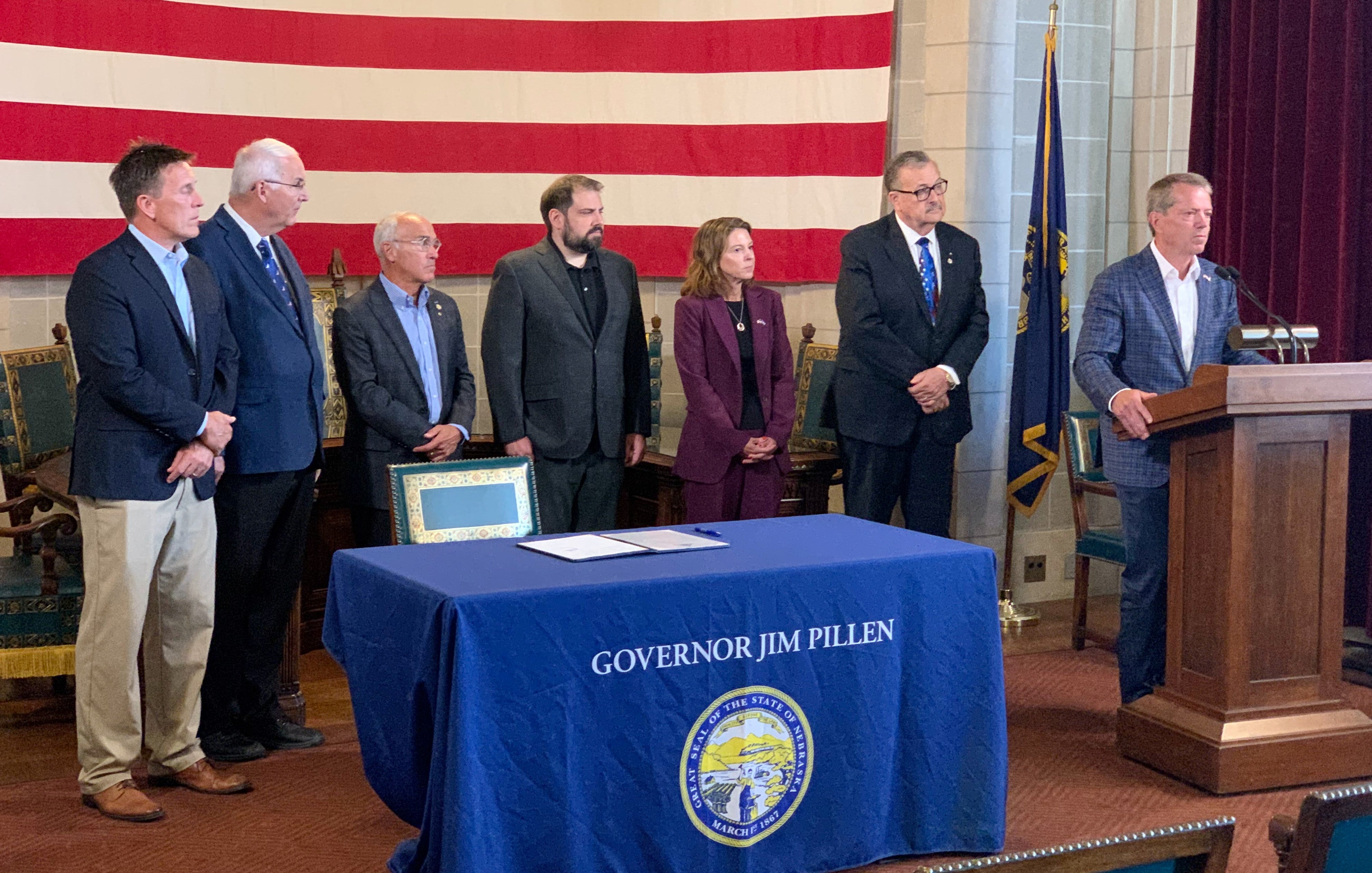 Gov. Pillen speaks from a podium at data center news conference. Joining him are Senator Mike Jacobson, Maureen Larsen, Director of Economic Development (DED), Director Jesse Bradley of Water, Energy and Environment (DWEE), and three other unidentified people. In front of the group is a table with a table cloth saying "Governor Jim Pillen" with the Great Seal of Nebraska under that. A huge American flag is hanging on the wall behind them.