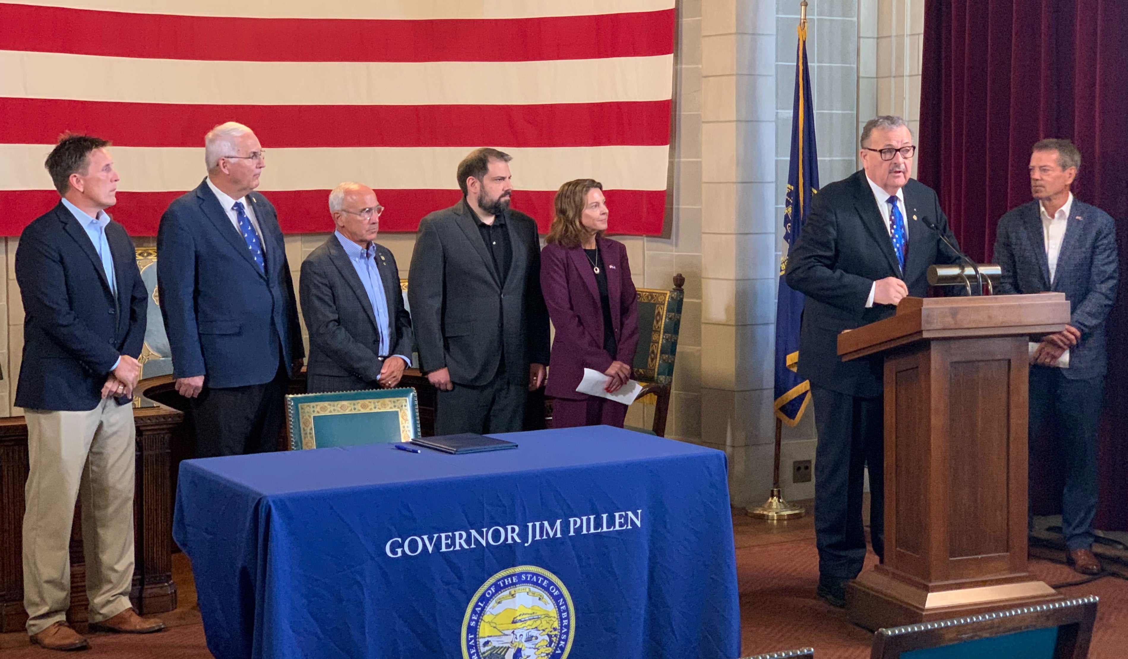 Senator Mike Jacobson speaks from a podium at data center news conference. Joining him are Gov. Pillen, Maureen Larsen, Director of Economic Development (DED), Director Jesse Bradley of Water, Energy and Environment (DWEE), and three other unidentified people. In front of the group is a table with a table cloth saying "Governor Jim Pillen" with the Great Seal of Nebraska under that. A huge American flag is hanging on the wall behind them.