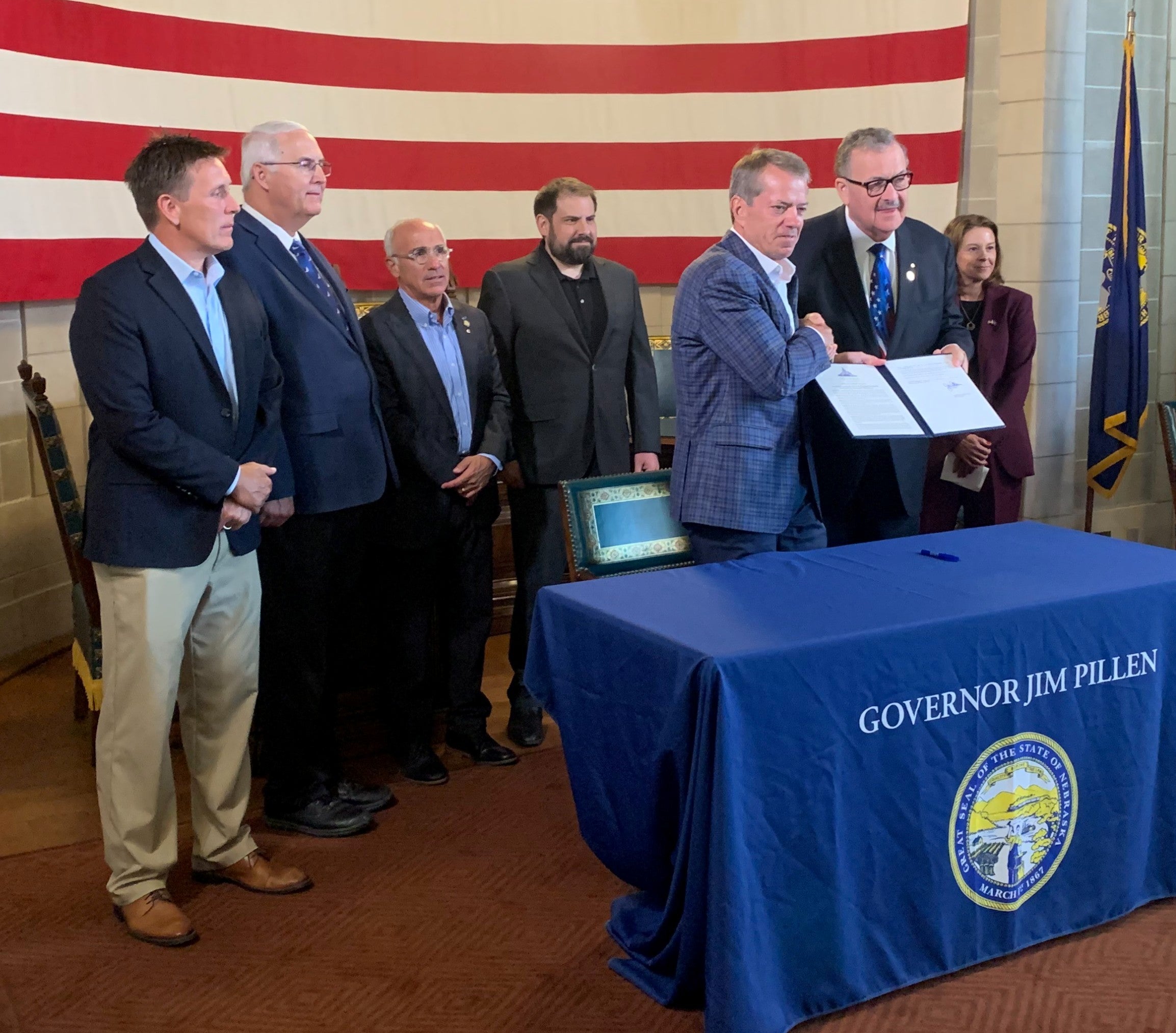 Gov. Pillen and Senator Mike Jacobson display signed executive order. Joining them are Maureen Larsen, Director of Economic Development (DED), Director Jesse Bradley of Water, Energy and Environment (DWEE), and three other unidentified people. In front of the group is a table with a table cloth saying "Governor Jim Pillen" with the Great Seal of Nebraska under that. A huge American flag is hanging on the wall behind them.