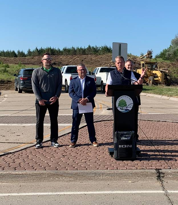 Governor Jim Pillen addresses local leaders, housing advocates and contractors in Blair at the ceremonial bill signing of LB 1114. Governor Pillen is standing behind a podium with a "City of Blair, Nebraska" emblem with a tree, stream, and bridge on it. A speaker is in front. He is joined by Senator Mike Jacobson, Adam Flanagan, and Jason Thiellen. In the background are an SUV, two pickups, a road grader, and evergreen trees.