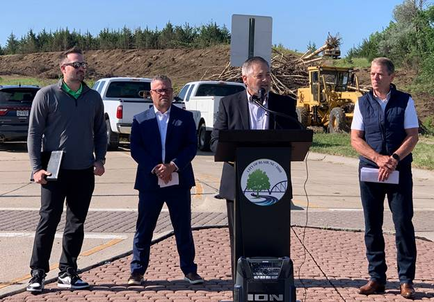 Adam Flanagan addresses local leaders, housing advocates and contractors in Blair behind the same podium and background as the Governor. He is joined by Senator Mike Jacobson, Jason Thiellen, and Governor Pillen. 