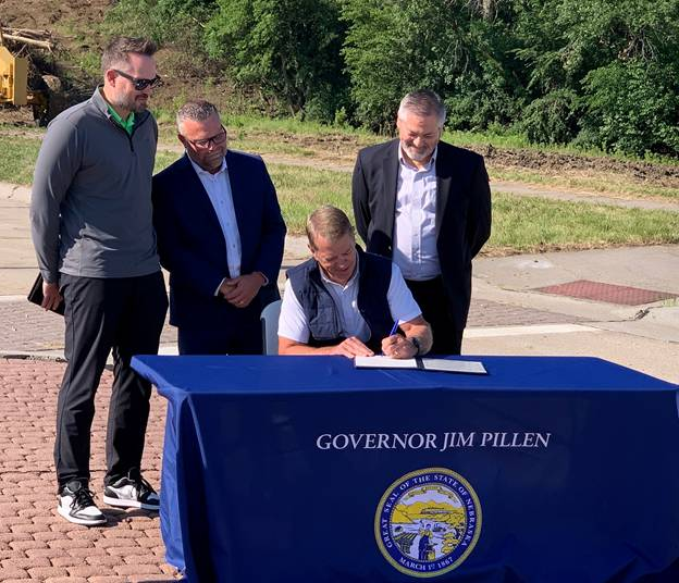 Governor Jim Pillen, sitting behind a table with a blue table cloth with "Governor Jim Pillen" written above an image of the Great Seal of the State of Nebraska, signs LB 1114 as Senator Mike Jacobson, Adam Flanagan, and Jason Thiellen look on.