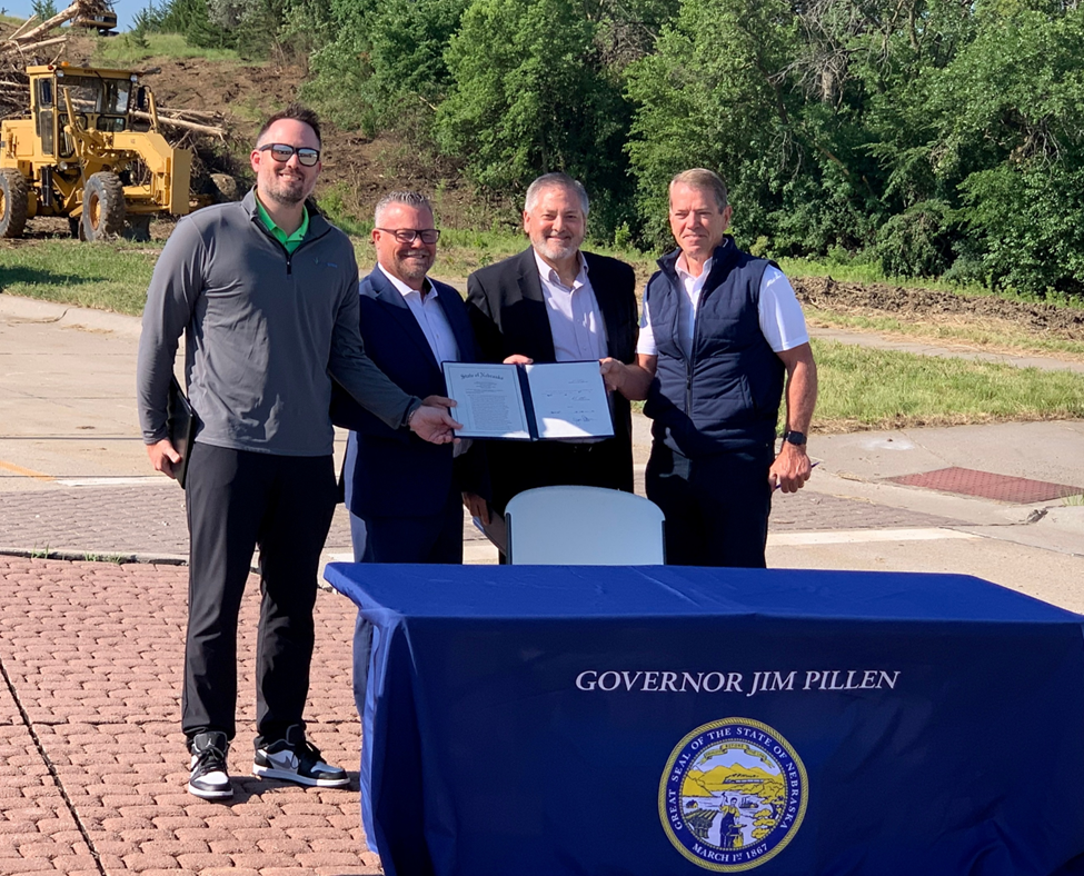 Senator Mike Jacobson, Adam Flanagan, Jason Thiellen and Governor Jim Pillen hold the signed bill while standing behind the table it was signed at.