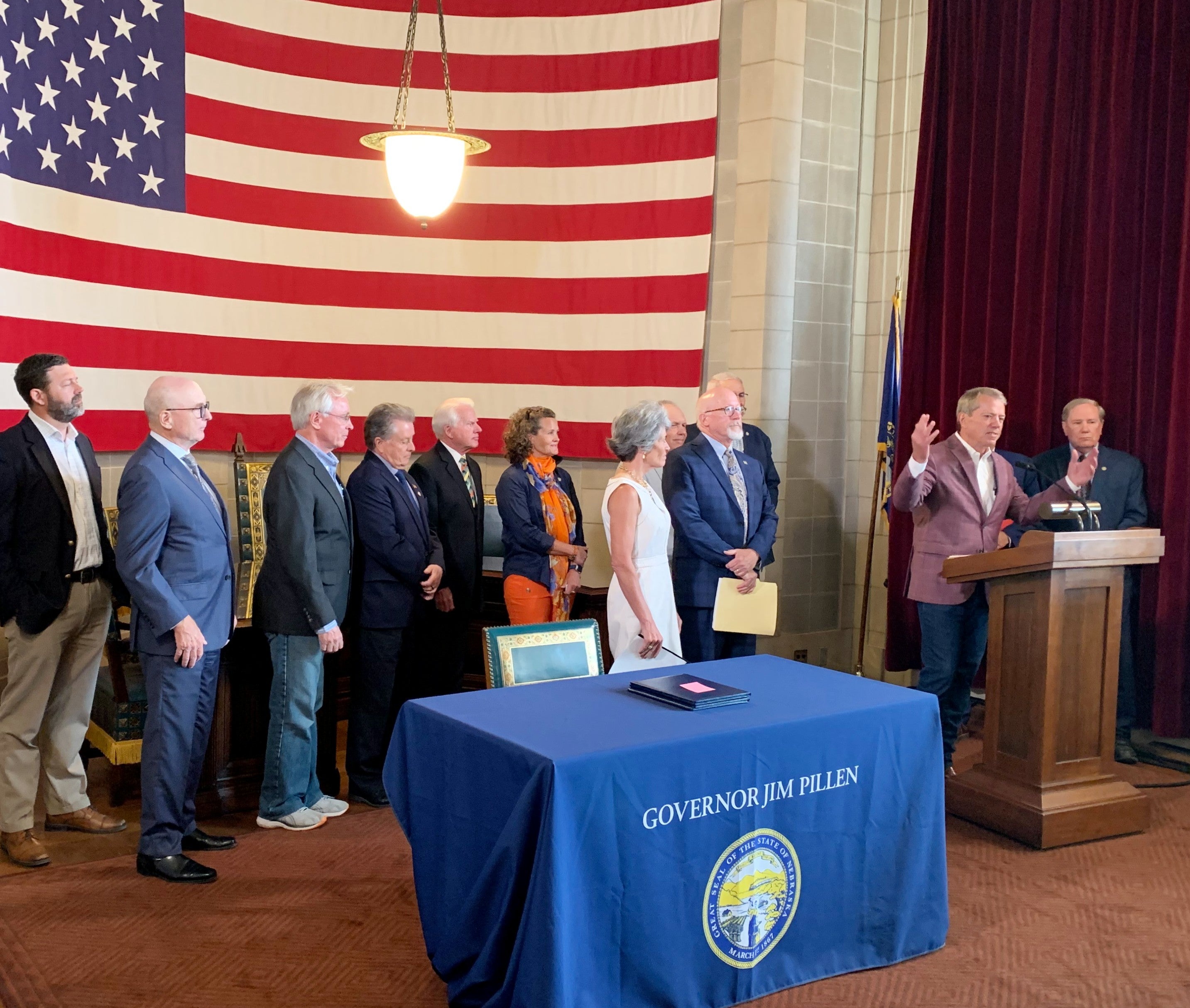 Governor Jim Pillen surrounded by state senators and representatives from public power entities addresses the audience from behind a podium. A table is in the foreground with a giant US flag hanging on the back wall.