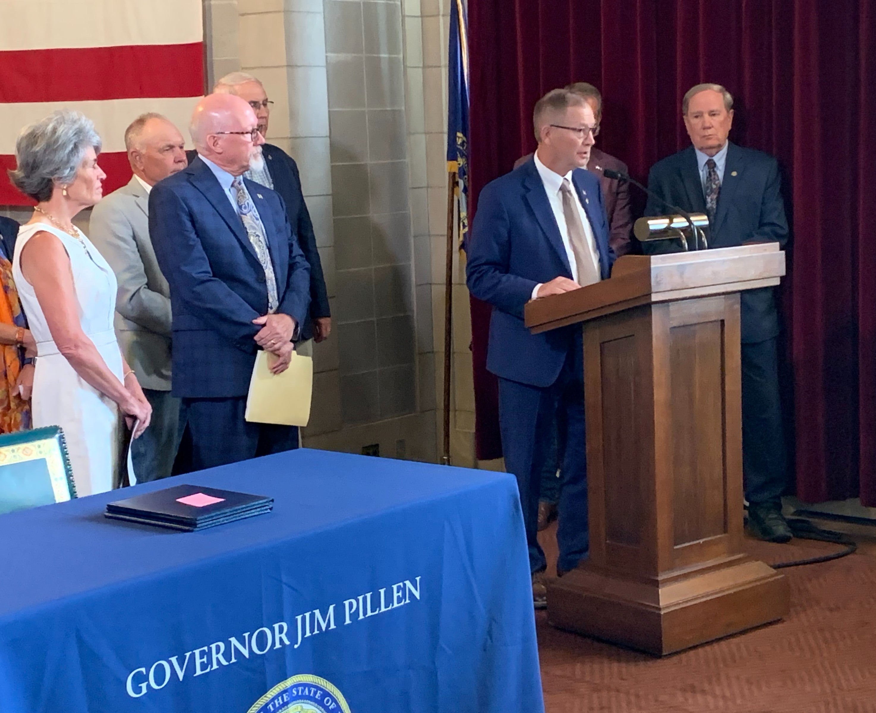 Senator Barry DeKay surrounded by the governor, state senators, and representatives from public power entities addresses the audience from behind a podium. A table is in the foreground with a giant US flag hanging on the back wall.