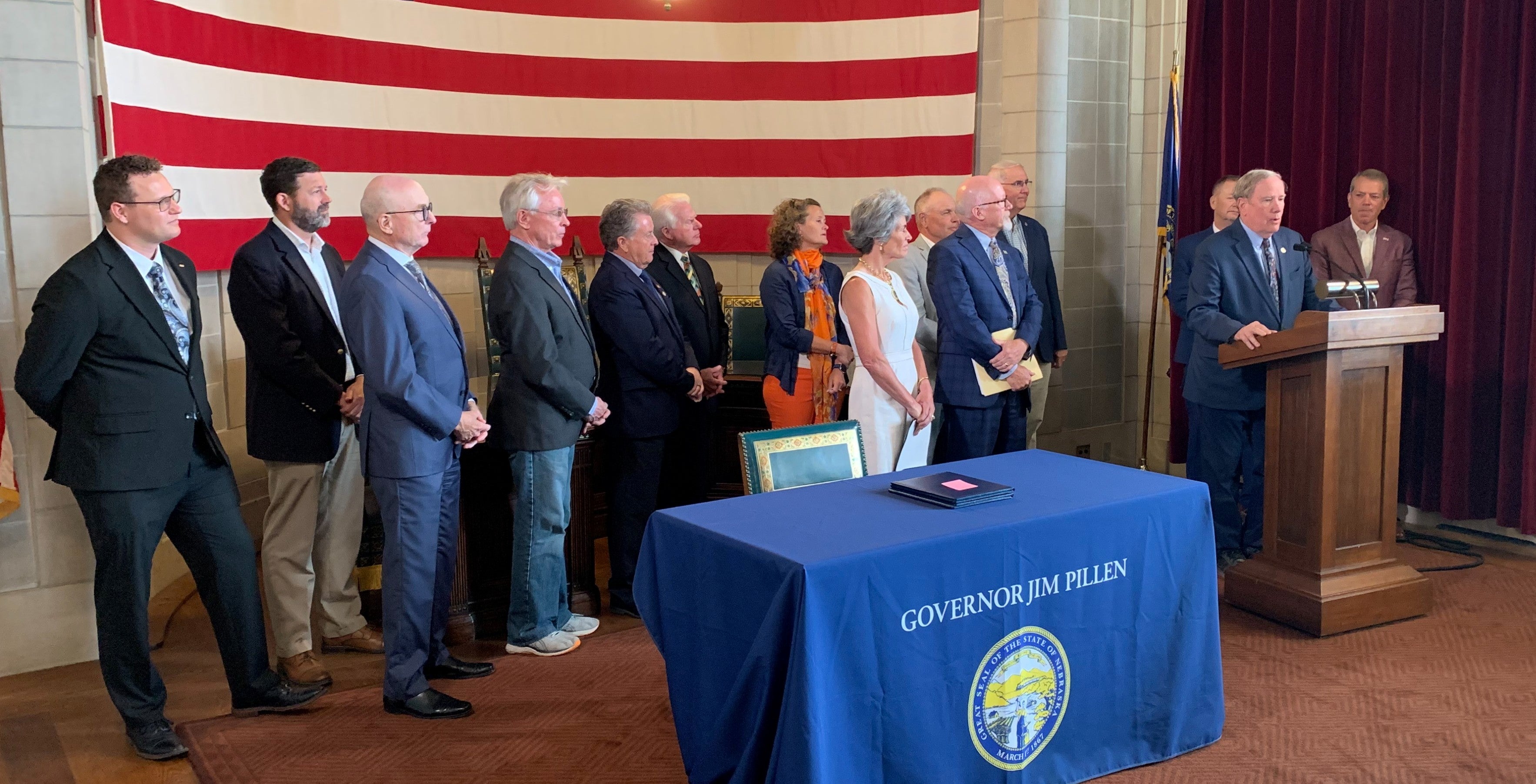 Senator Mike Moser surrounded by the governor, state senators, and representatives from public power entities addresses the audience from behind a podium. A table is in the foreground with a giant US flag hanging on the back wall.