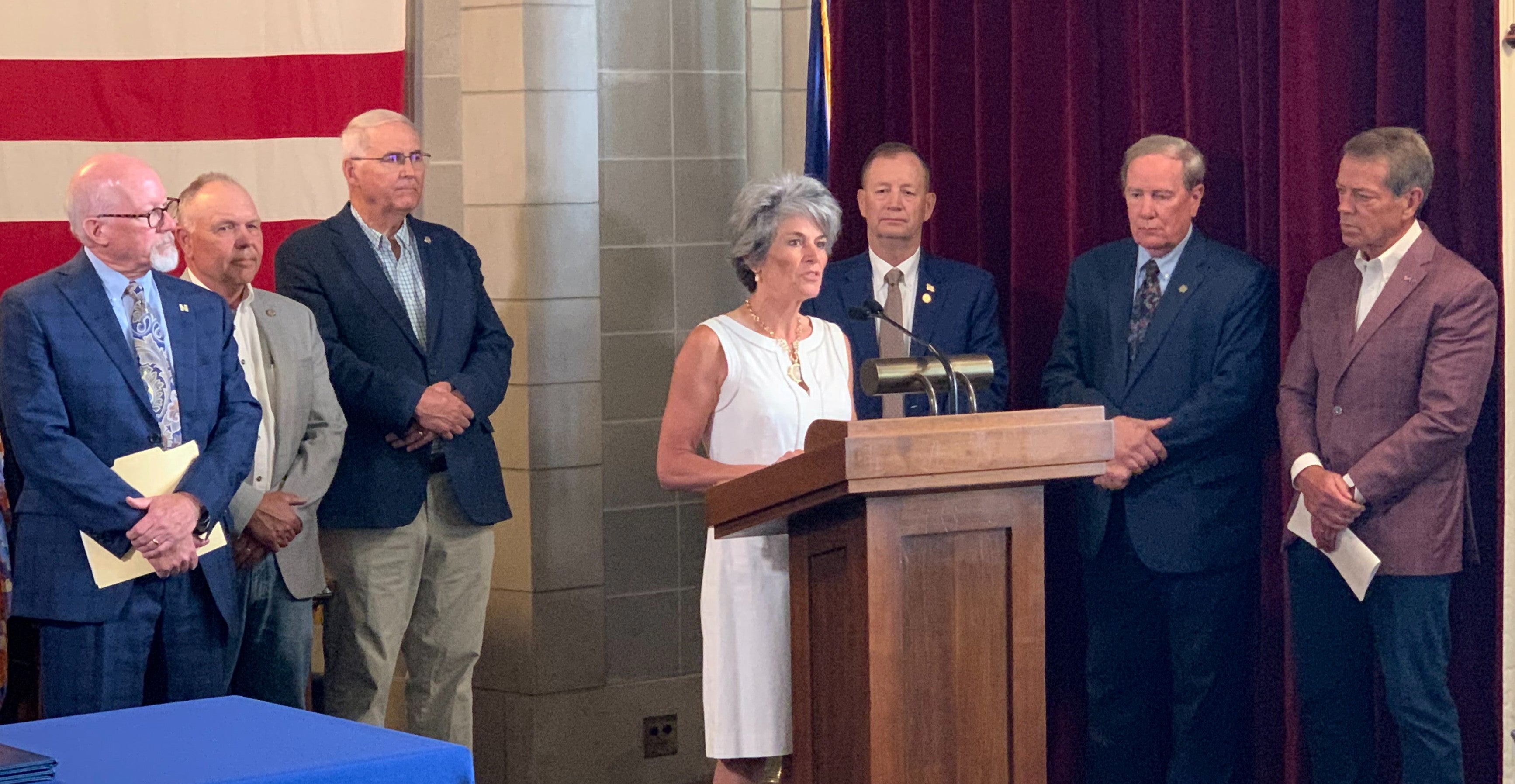 Speaking on behalf of Tenaska, Delette Marengo, surrounded by the governor, state senators, and representatives from public power entities, addresses the audience from behind a podium. A table is in the foreground with a giant US flag hanging on the back wall.