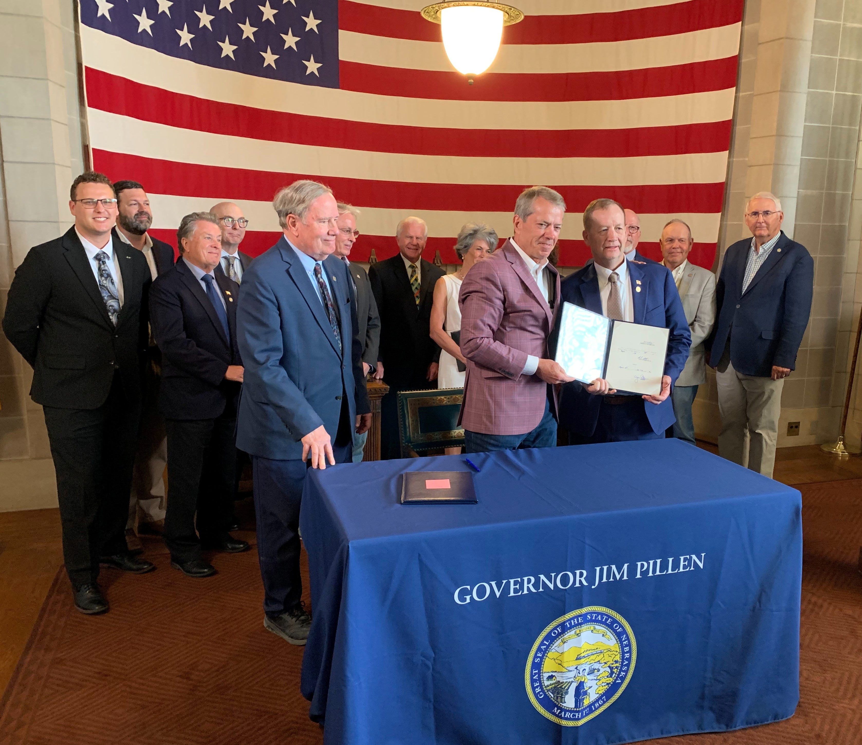 Governor Jim Pillen and Senator Barry DeKay, surrounded by state senators and representatives from public power entities, hold the signed bill behind the table with the giant US flag hanging on the back wall.