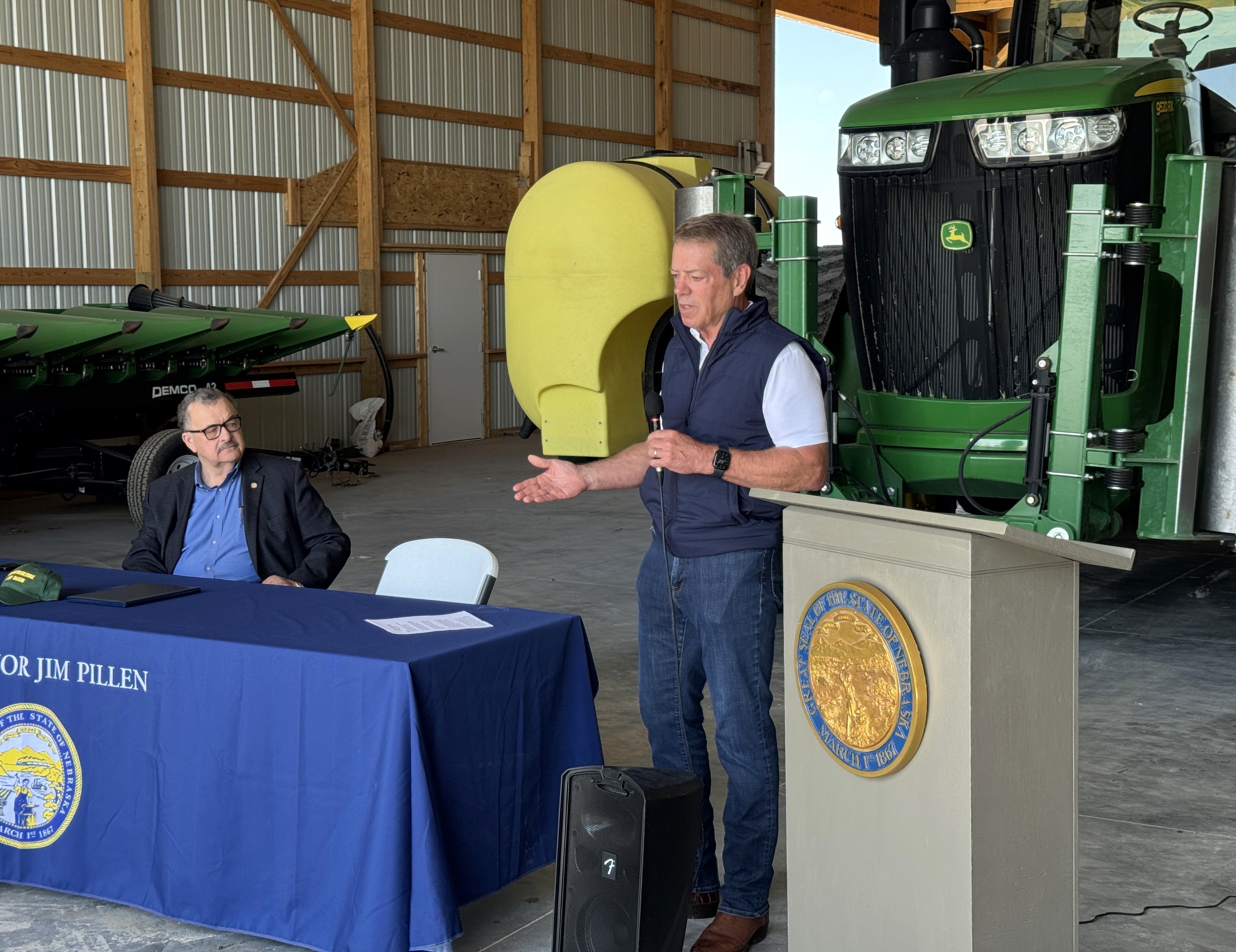 Governor Jim Pillen, standing between a podium and table covered with a cloth saying "Governor Jim Pillen", both with the Great Seal of Nebraska adorning them, addresses members of Nebraska’s agricultural industry at the Machine Shed near Gothenburg. Senator Mike Jacobson looks on from his seat. Tractor equipment sits in the background.