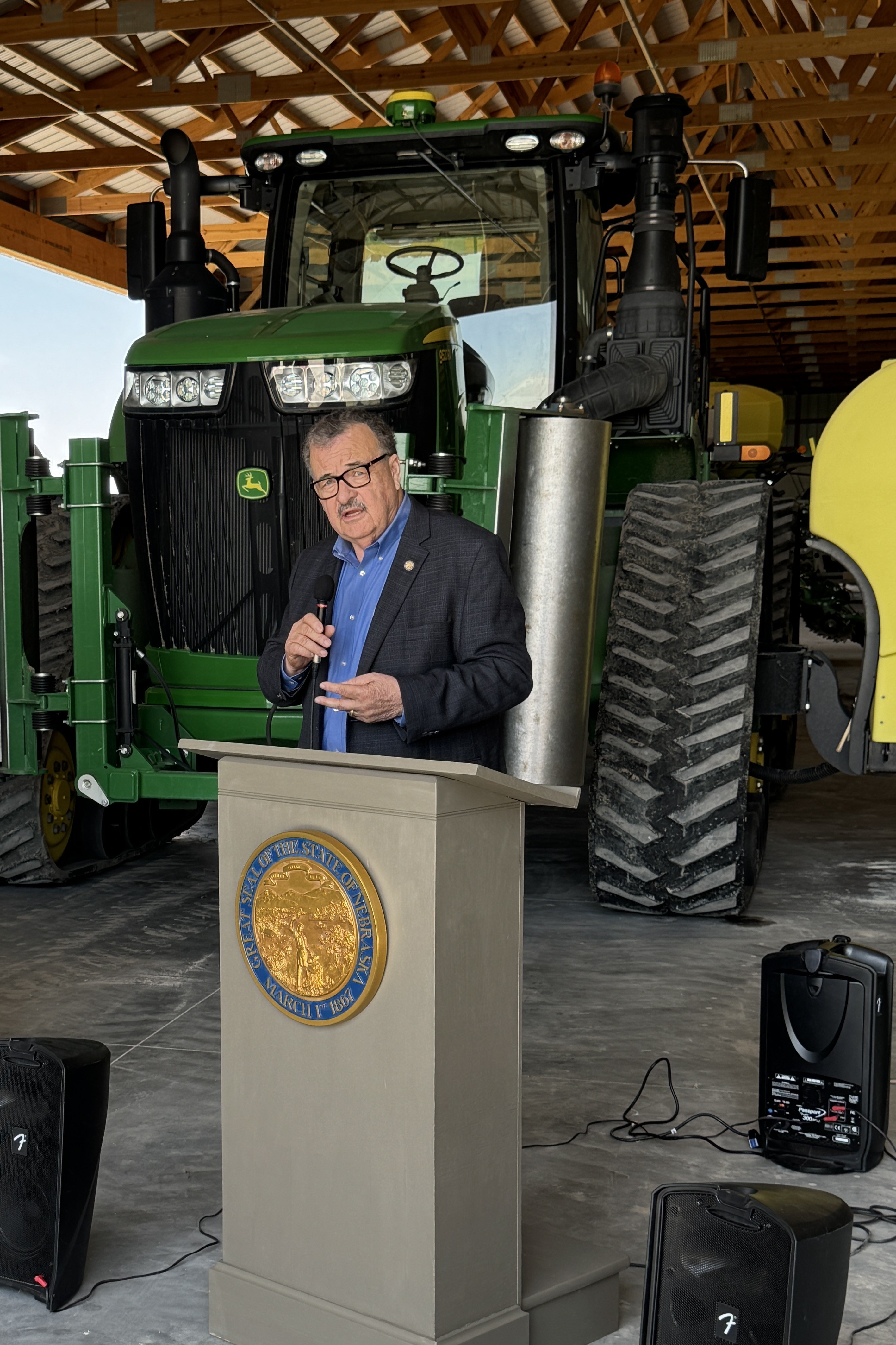 Senator Mike Jacobson, standing behind a podium with the Great Seal of Nebraska adorning it and speaker equipment on both sides, addresses members of Nebraska’s agricultural industry at the Machine Shed near Gothenburg. A John Deer tractor is in the background.