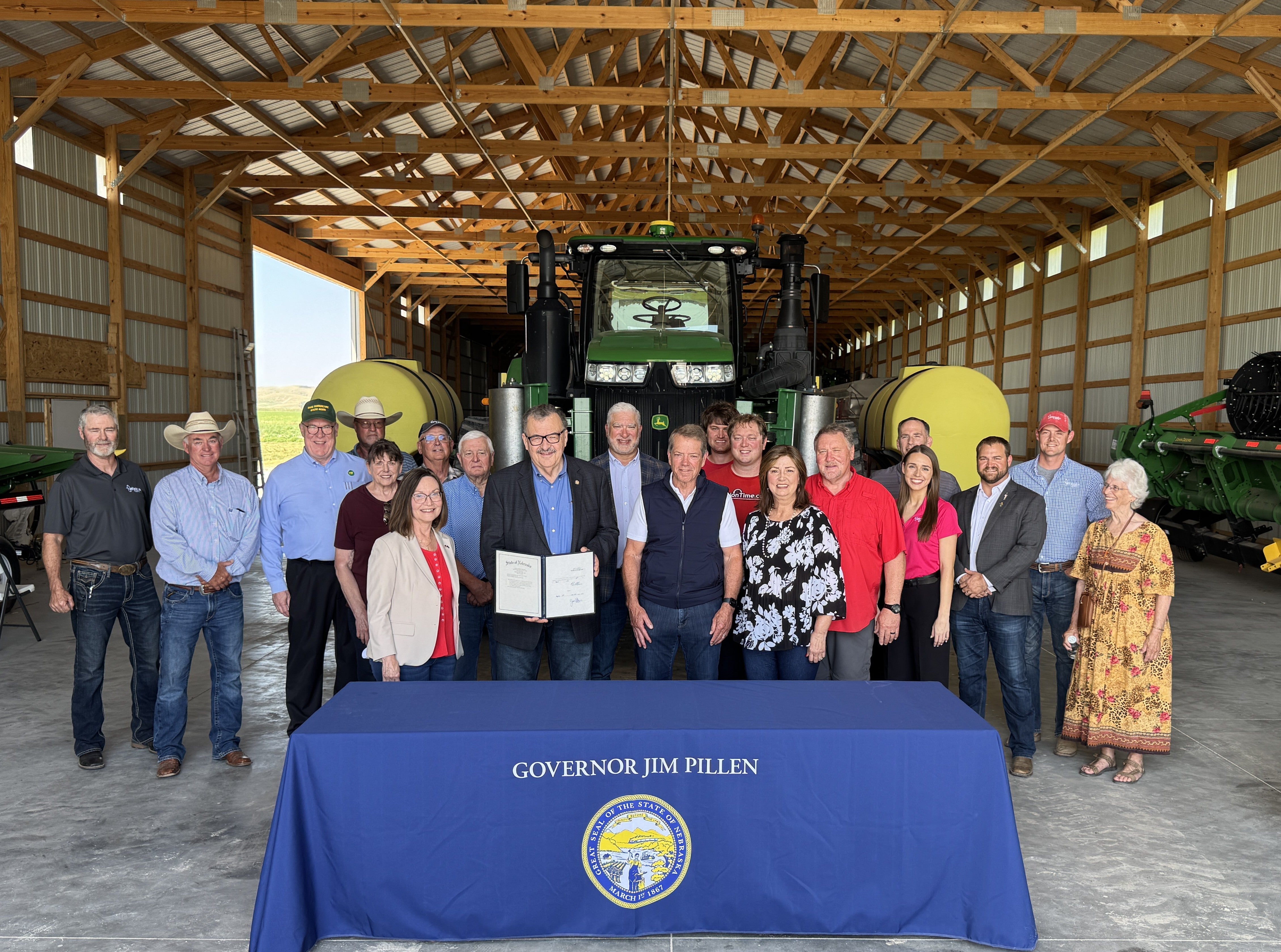 Group photo of Governor Jim Pillen, Senator Mike Jacobson, and members of Nebraska’s agricultural industry at the Machine Shed near Gothenburg, stand behind a table with a cloth saying "Governor Jim Pillen" on it. John Deer tractor equipment is in the background, as well as an open door showing the countryside.