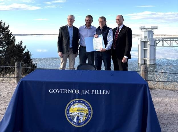 Standing behind a table with a blue tablecloth with Governor Jim Pillen written above the Great Seal of the State of Nebraska, Governor Jim Pillen and the team were joined by Game and Parks Director Tim McCoy at the edge of Lake McConaughy displaying a signed copy of a program statement that promises $16 million in appropriations to the agency for infrastructure development.
