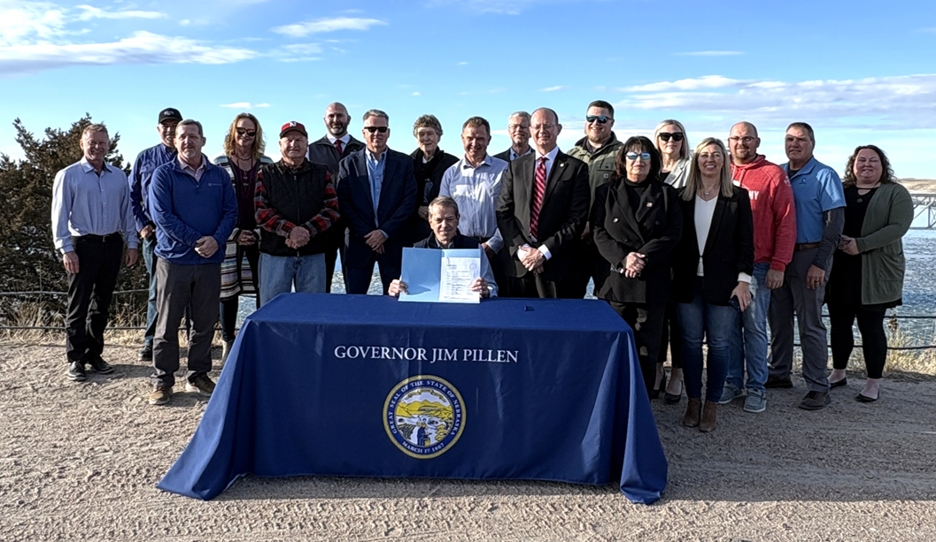 Sitting behind a table with a blue tablecloth with Governor Jim Pillen written above the Great Seal of the State of Nebraska, Governor Jim Pillen and the team were joined by Game and Parks Director Tim McCoy and a plethora of representatives from Ogallala, Nebraska at the edge of Lake McConaughy displaying a signed copy of a program statement that promises $16 million in appropriations to the agency for infrastructure development.