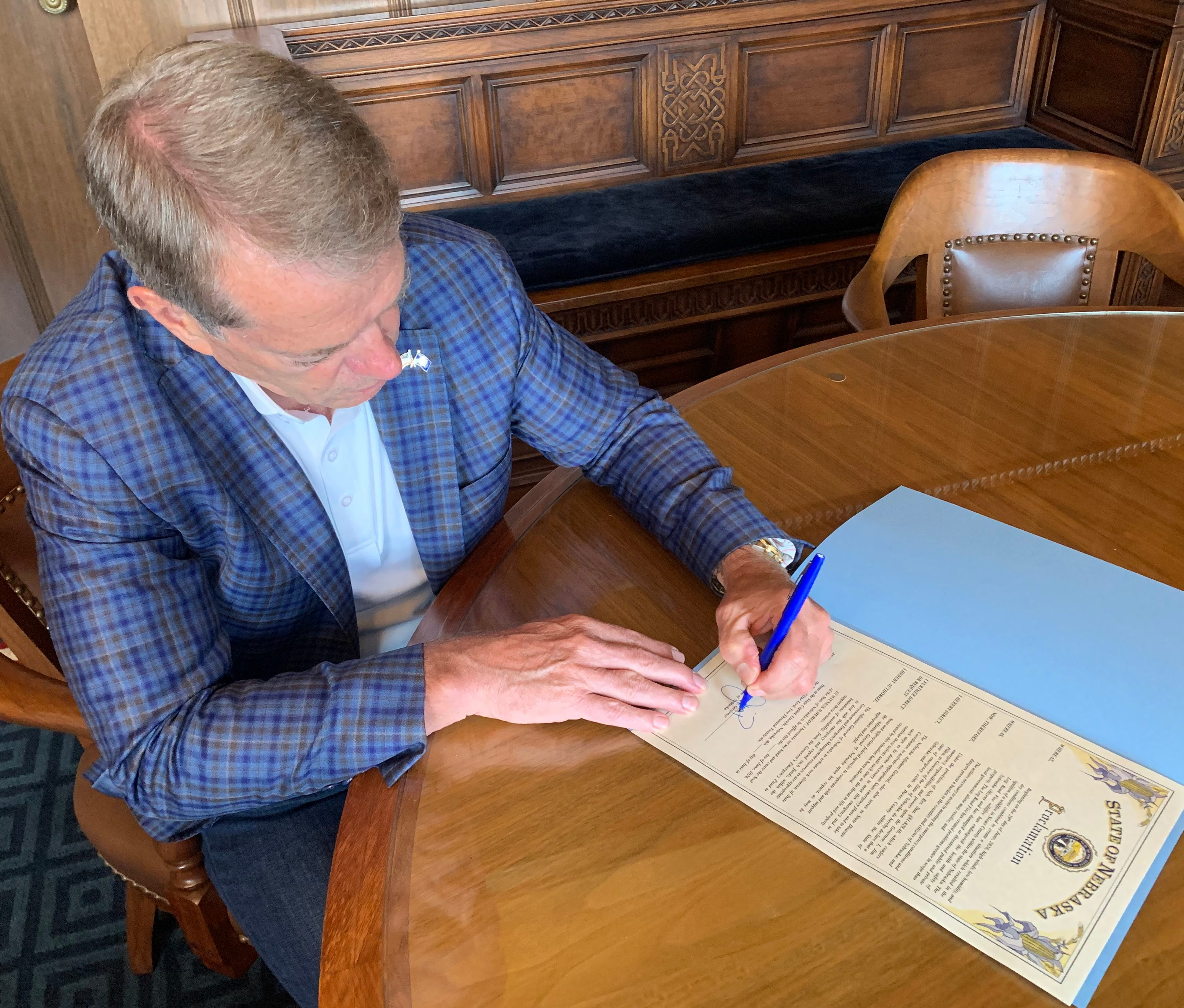 Governor Jim Pillen signs the Log Road Fire Proclamation while seated at a round wooden table.