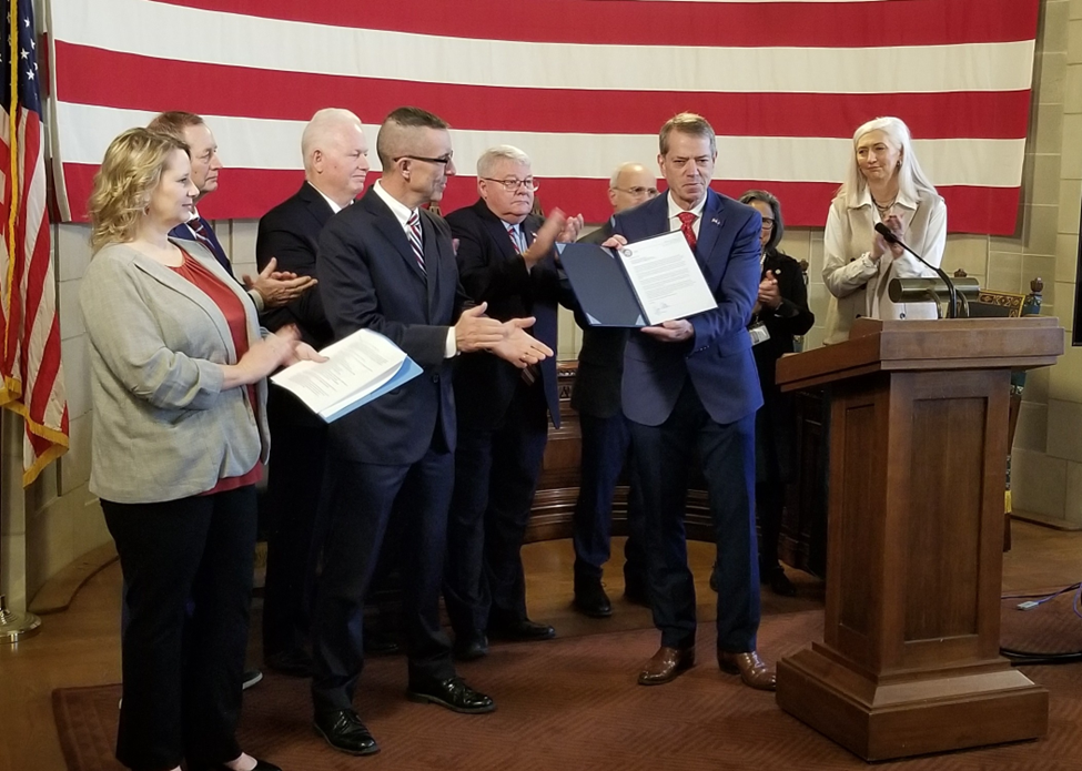 Governor Pillen holds the signed order while State leaders applaud.