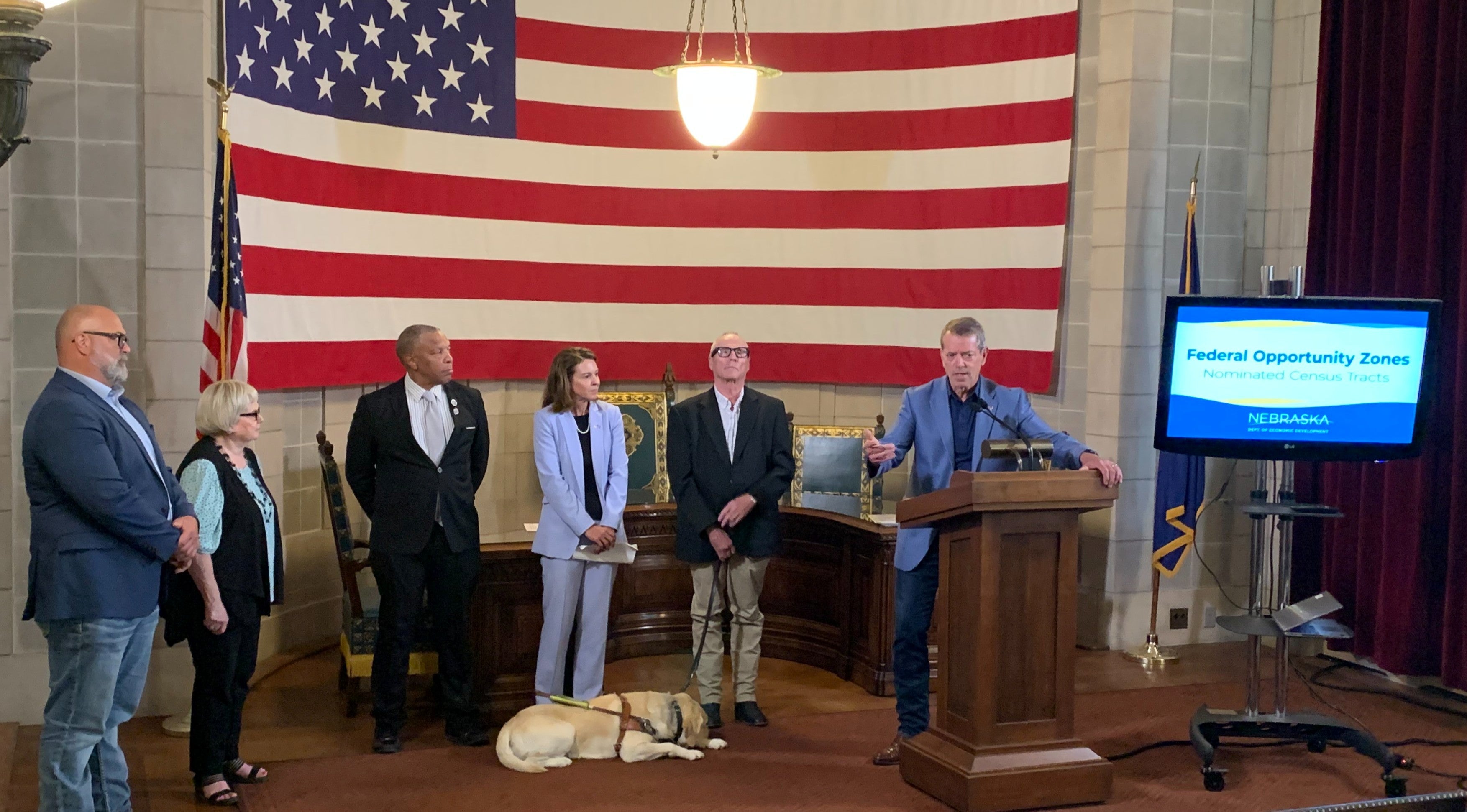 Gov. Pillen addresses attendees of Federal Opportunity Zones announcement. The Governor was joined for the announcement by Nebraska Department of Economic Development (DED) Director Maureen Larsen, Omaha Mayor John Ewing, Lexington Mayor John Fagot, McCook Mayor Linda Taylor, and Norfolk Mayor Shane Clausen. In the picture are a service dog, a television monitor with "Federal Opportunity Zones" displayed, and a giant American flag in the background.
