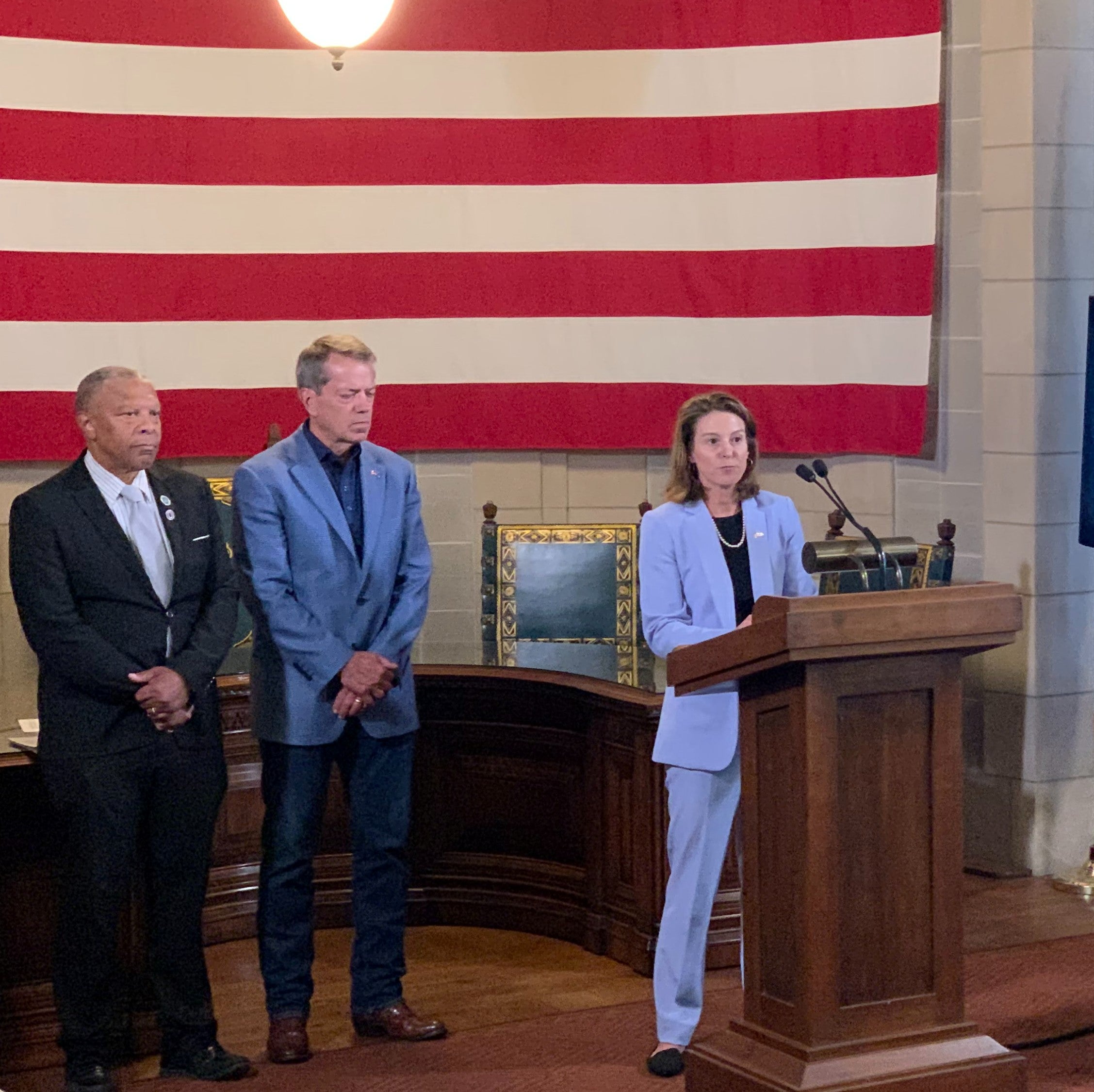 DED Director Maureen Larsen addresses attendees of Federal Opportunity Zones announcement. Governor Pillen and Mayer Ewing stand beside her with the giant American flag as a backdrop.