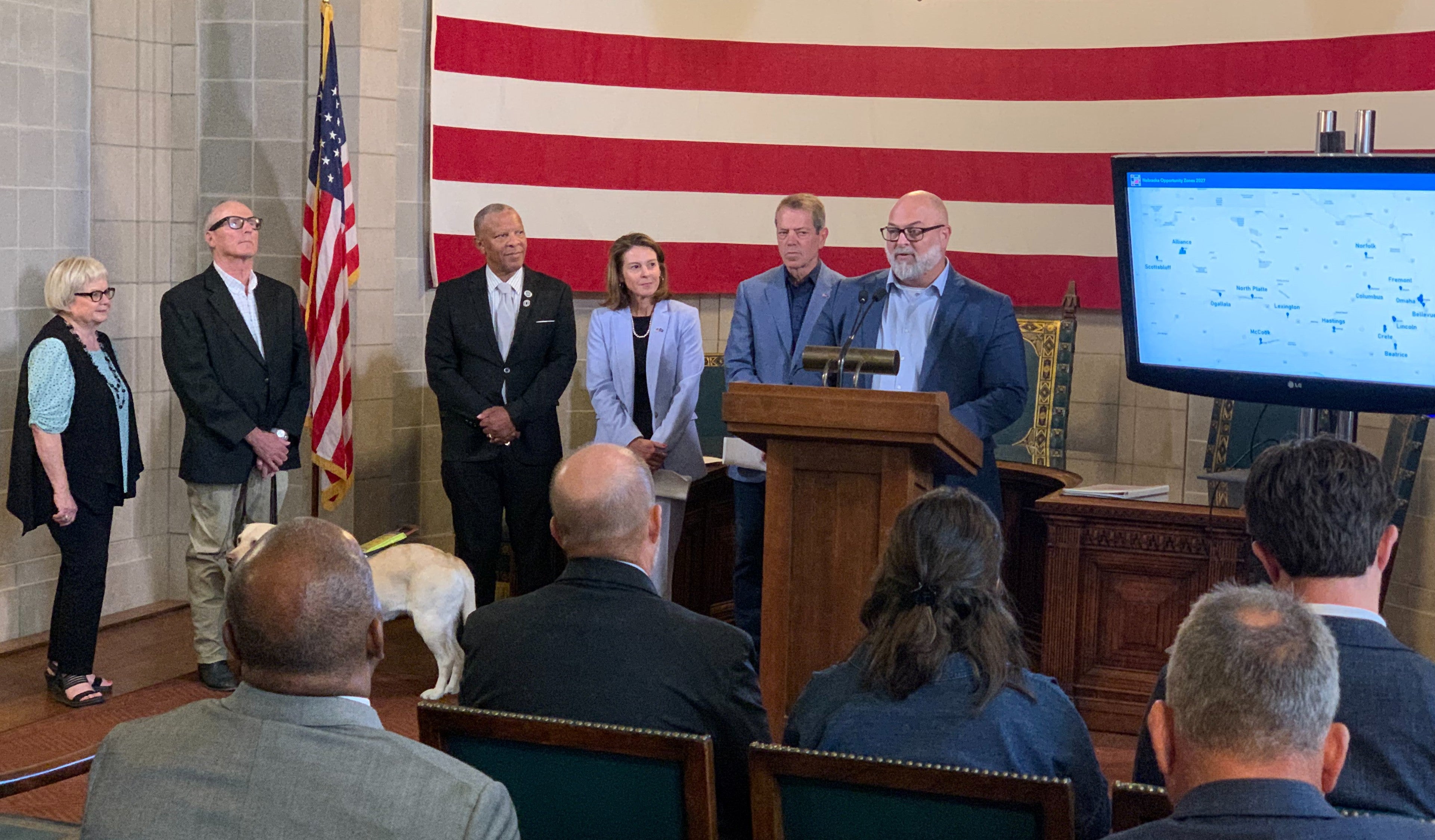 Norfolk Mayor Shane Clausen addresses attendees of Federal Opportunity Zones announcement. was joined for the announcement by Nebraska Department of Economic Development (DED) Director Maureen Larsen, Omaha Mayor John Ewing, Lexington Mayor John Fagot, McCook Mayor Linda Taylor, and Norfolk Mayor Shane Clausen. In the picture are Governonr Pillen, audience members, a service dog, a television monitor with a map of Nebraska showing the selected zones is displayed, and a giant American flag in the background.