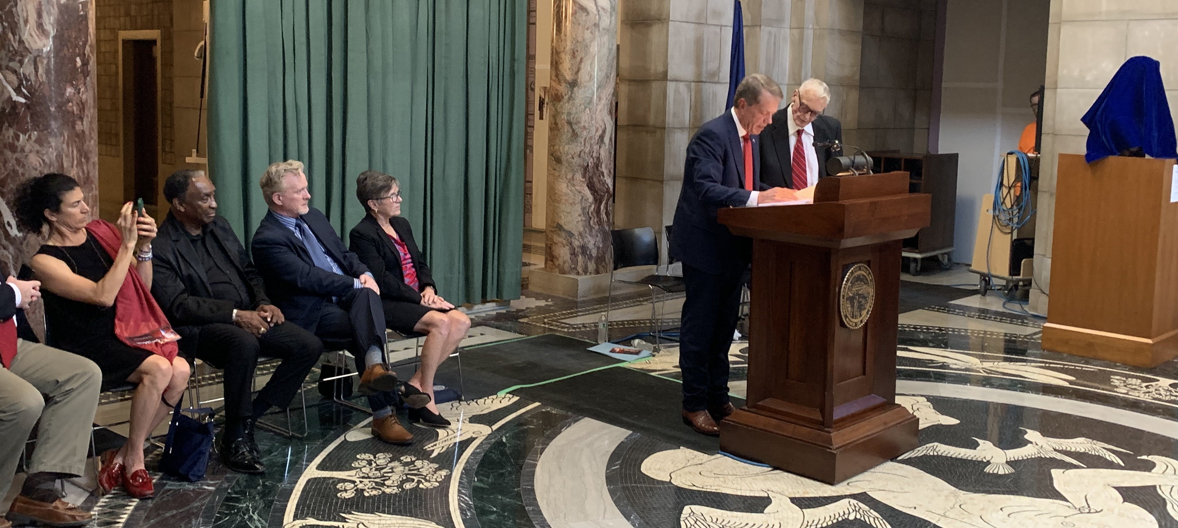 Gov. Pillen, standing at a podium with Coach Tom Osborne, reads the proclamation  marking Coach Tom Osborne Hall of Fame Day as members of the Hall of Fame ommittee look on