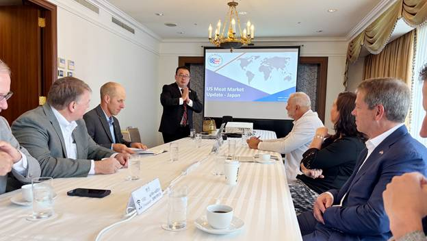 Gov. Pillen and delegation members meet with U.S. Meat Export Federation representatives in Japan. They are seated at a table with a white table cloth with water glasses and coffee cups on the table. In the background is a screen with a map of the world and US Meat Market Update - Japan displayed.