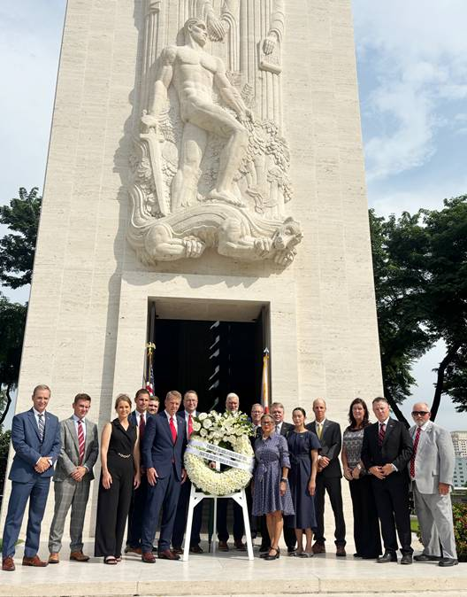 Gov. Pillen and unnamed delegation members at Manila American Cemetery and Memorial. There is a structure in the background with a doorway flanked by a USA flag and another unknown flag. Above the doorway on the structure is a design of a warrior holding a sword standing on a dragon that appears to be dead. Two figures stand behind the warrior.