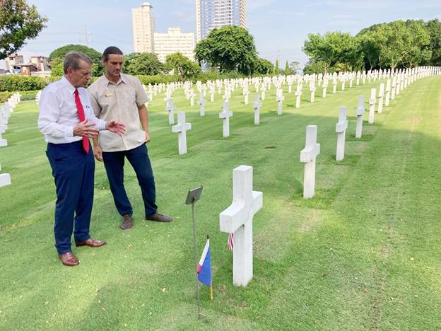 Gov. Pillen visits Manila American Cemetery and Memorial. Gov. Pillen talks with an unnamed man with rows of white crosses used as grave markers in a grass covered field. Trees and high rise buildings stand in the background.