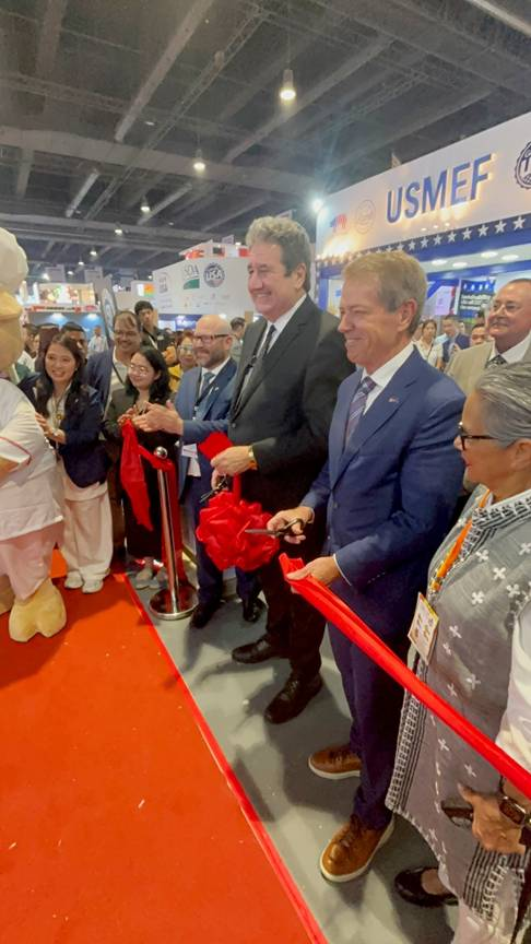 With a crowd gathered around, Gov. Pillen cuts a red ribbon at a booth to promote U.S. products during the World Food Expo while U.S. Ambassador Lee Lipton, Senator Sanders, and other unnamed delegation members watch.