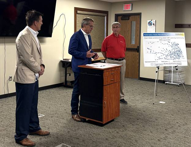 Nebraska Department of Water, Energy, and Environment (DWEE) Director Jesse Bradley, Governor Jim Pillen, and Plainview Mayor Bob Smith stand in a meeting room during a presentation, with Governor Pillen speaking at a podium. A display board behind them shows a map and information about Nebraska state revolving funds for clean water and drinking water projects.