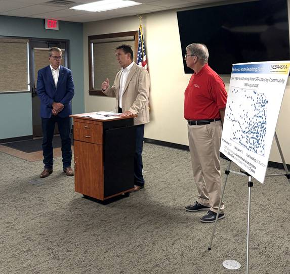 Governor Jim Pillen, Nebraska Department of Water, Energy, and Environment (DWEE) Director Jesse Bradley, and Plainview Mayor Bob Smith stand in a meeting room during a presentation, with Director Bradley speaking at a podium. An American flag and a display board showing Nebraska State Revolving Fund information and a map of project locations are visible in the background.