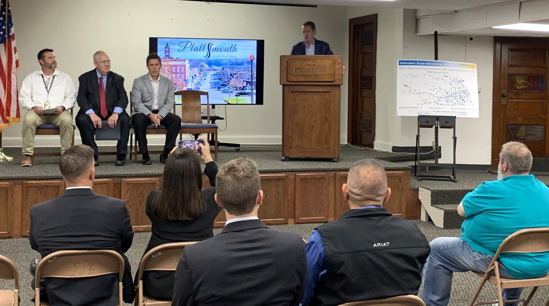 Governor Jim Pillen, speaking from a podium on a stage with a television screen in the background and a poster to his right, is joined on stage by, from left to right, NEMA Recovery Section Administrator Donny Christensen, Plattsmouth Mayor R. Paul Lambert, and Nebraska Department of Water, Energy, and Environment (DWEE) Director Jesse Bradley.