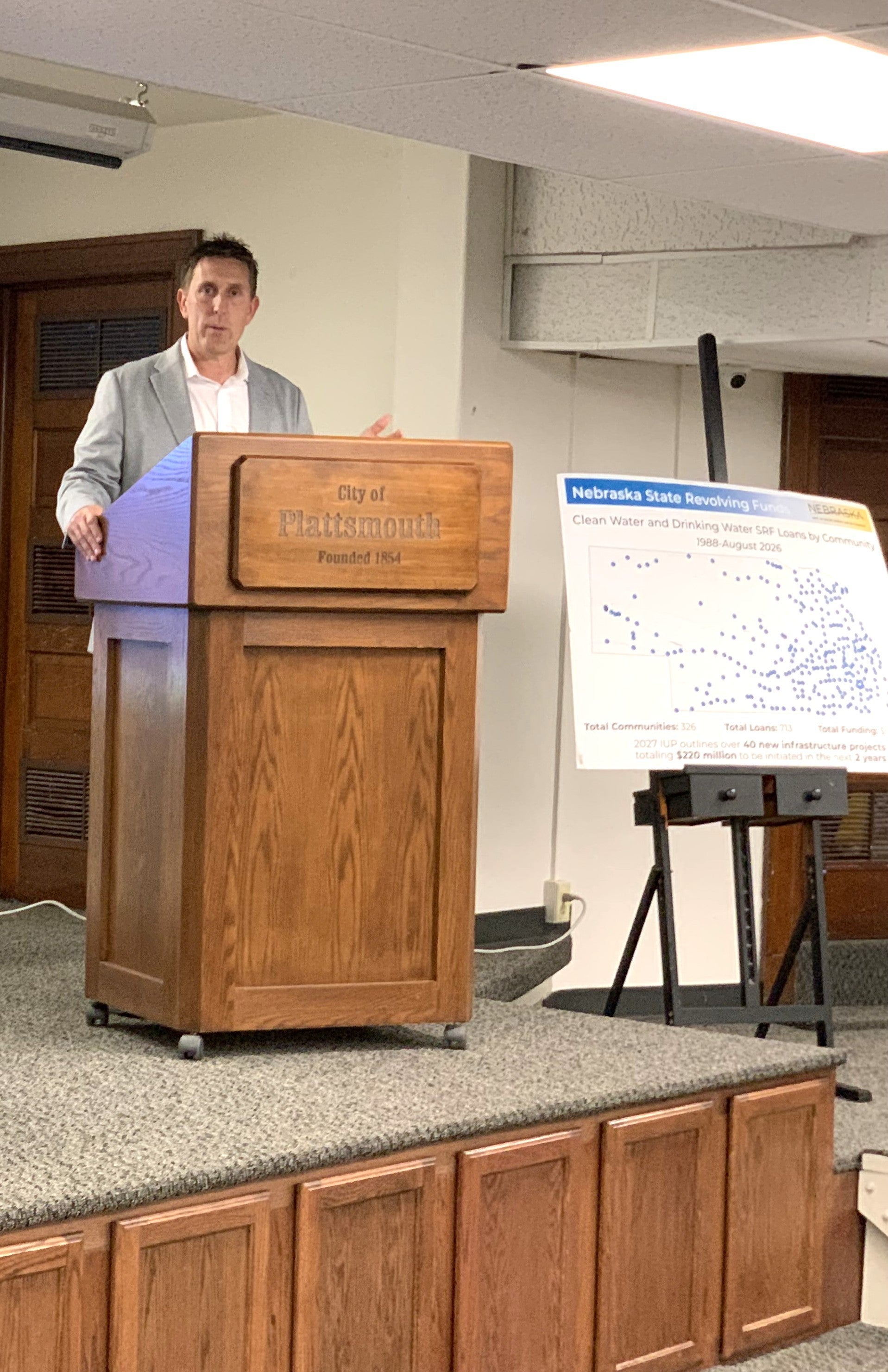 Nebraska Department of Water, Energy, and Environment (DWEE) Director Jesse Bradley speaks from the podium on the stage. Next to him is a poster titled "Nebraska State Revolving Fund" with a caption of "Clean Water and Drinking Water SRF Loans by Communities 1988-August 2026" with hundreds of blue dots representing the cities serviced.