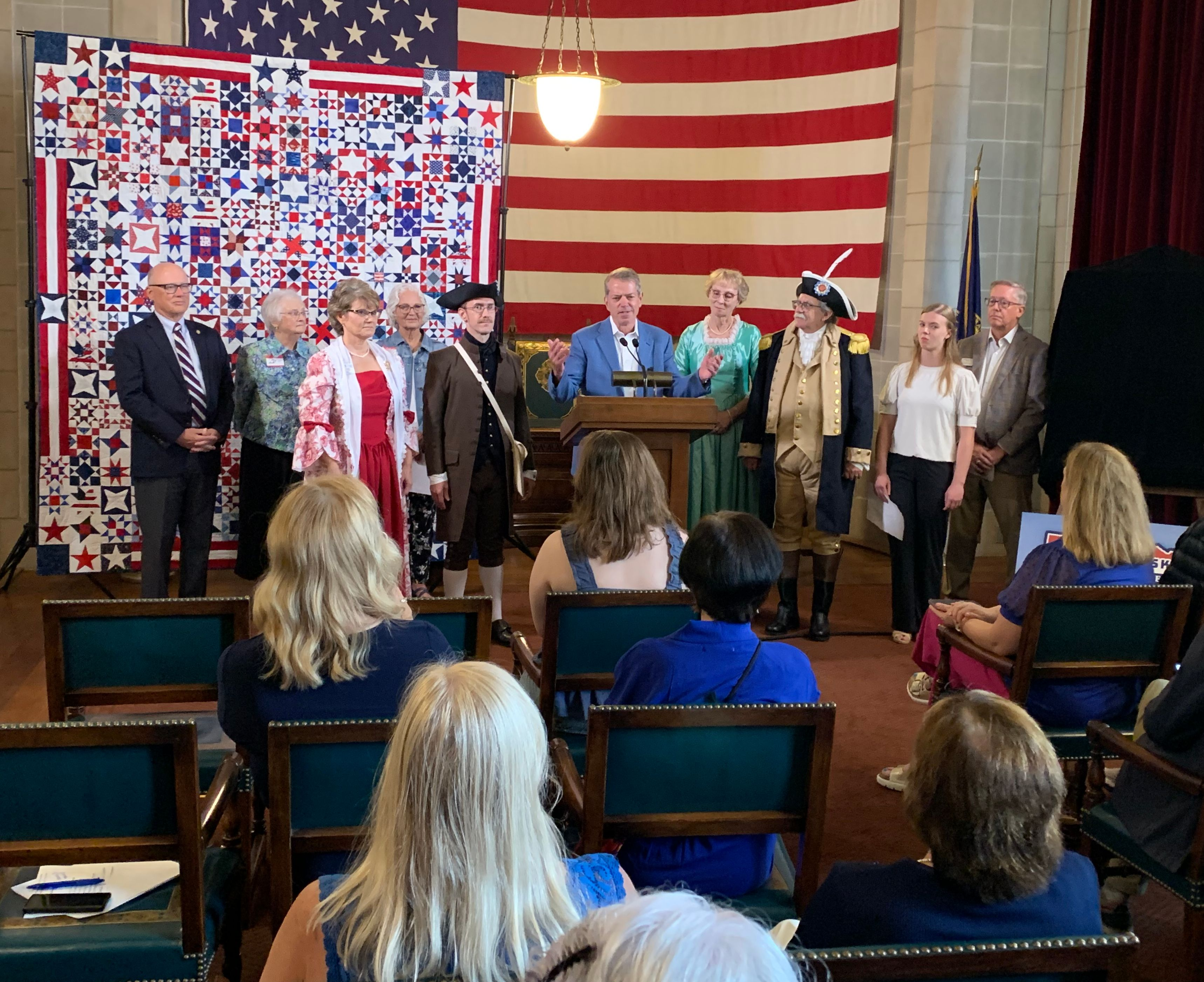 Governor Jim Pillen, flanked by members of the Sons and Daughters of the American Revolution, as well as others, speaks to an audience. The quilt, with 250-star blocks colored red, white, and blue and a huge American flag are in the background. 