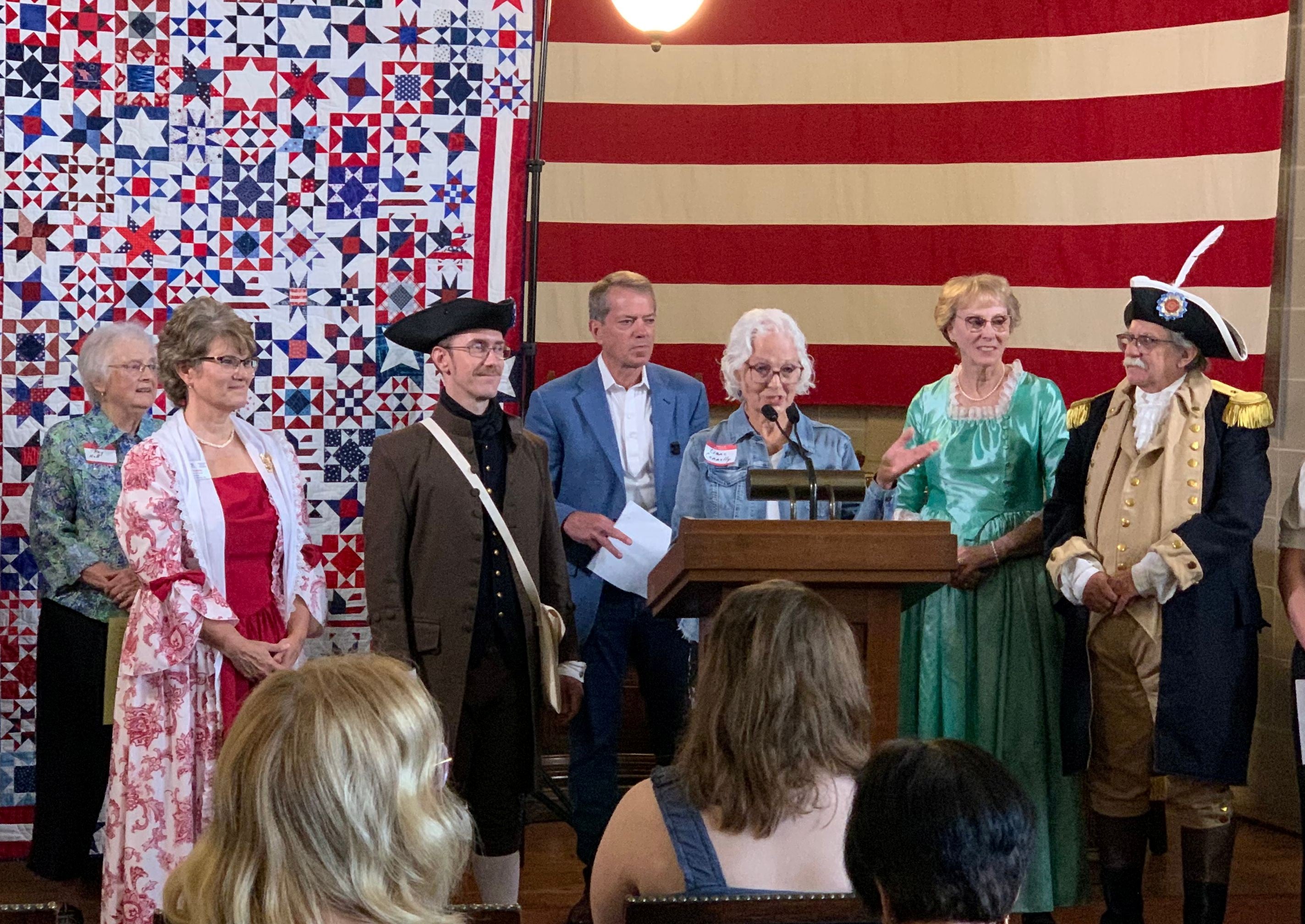 Lianne Connelly, president of the Lincoln Quilters Guild, flanked by Governor Jim Pillen, members of the Sons and Daughters of the American Revolution, as well as others, speaks to an audience. The quilt, with 250-star blocks colored red, white, and blue and a huge American flag are in the background. 
