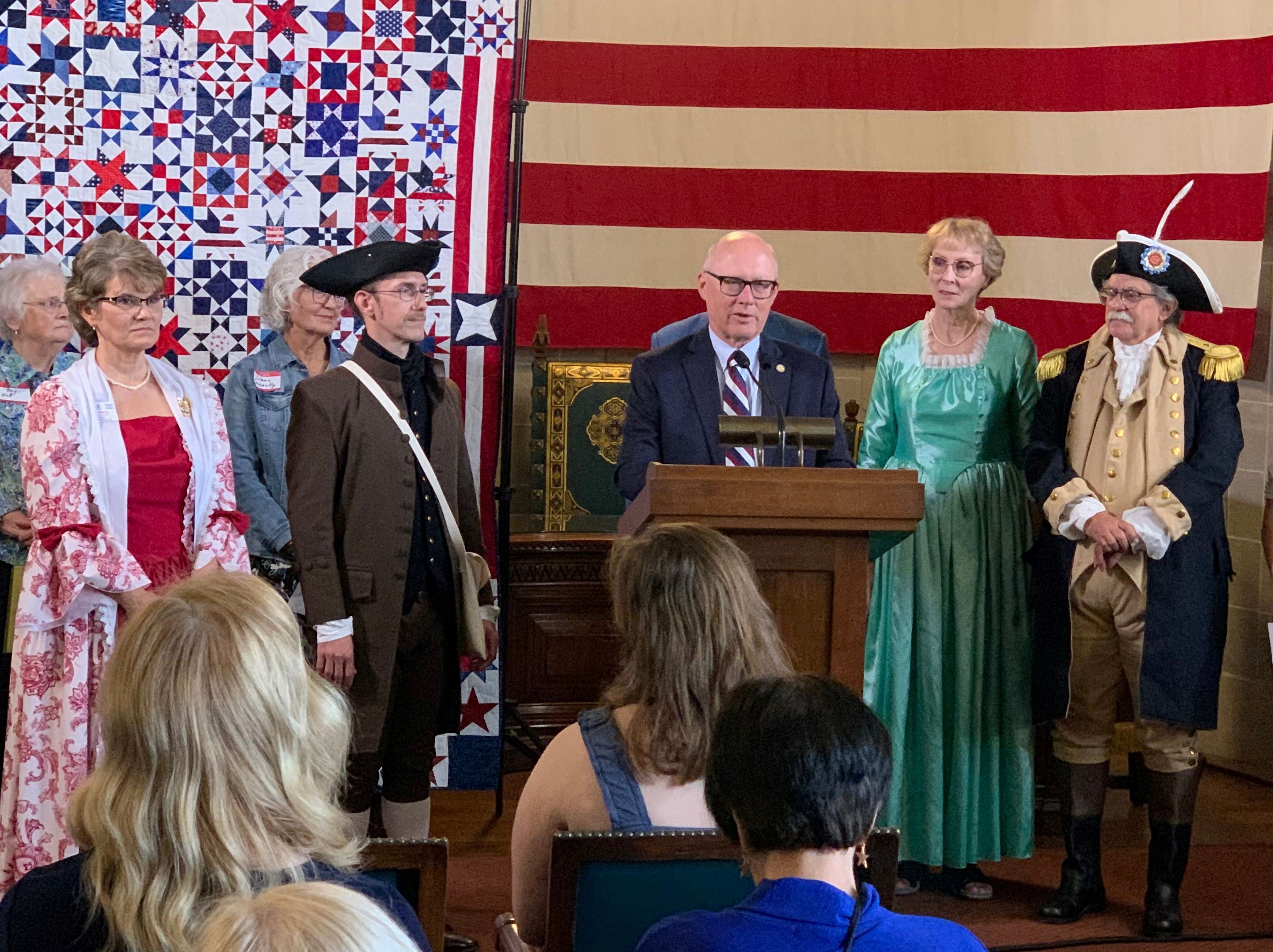 Senator John Arch, flanked by members of the Sons and Daughters of the American Revolution, as well as others, speaks to an audience. The quilt, with 250-star blocks colored red, white, and blue and a huge American flag are in the background. 