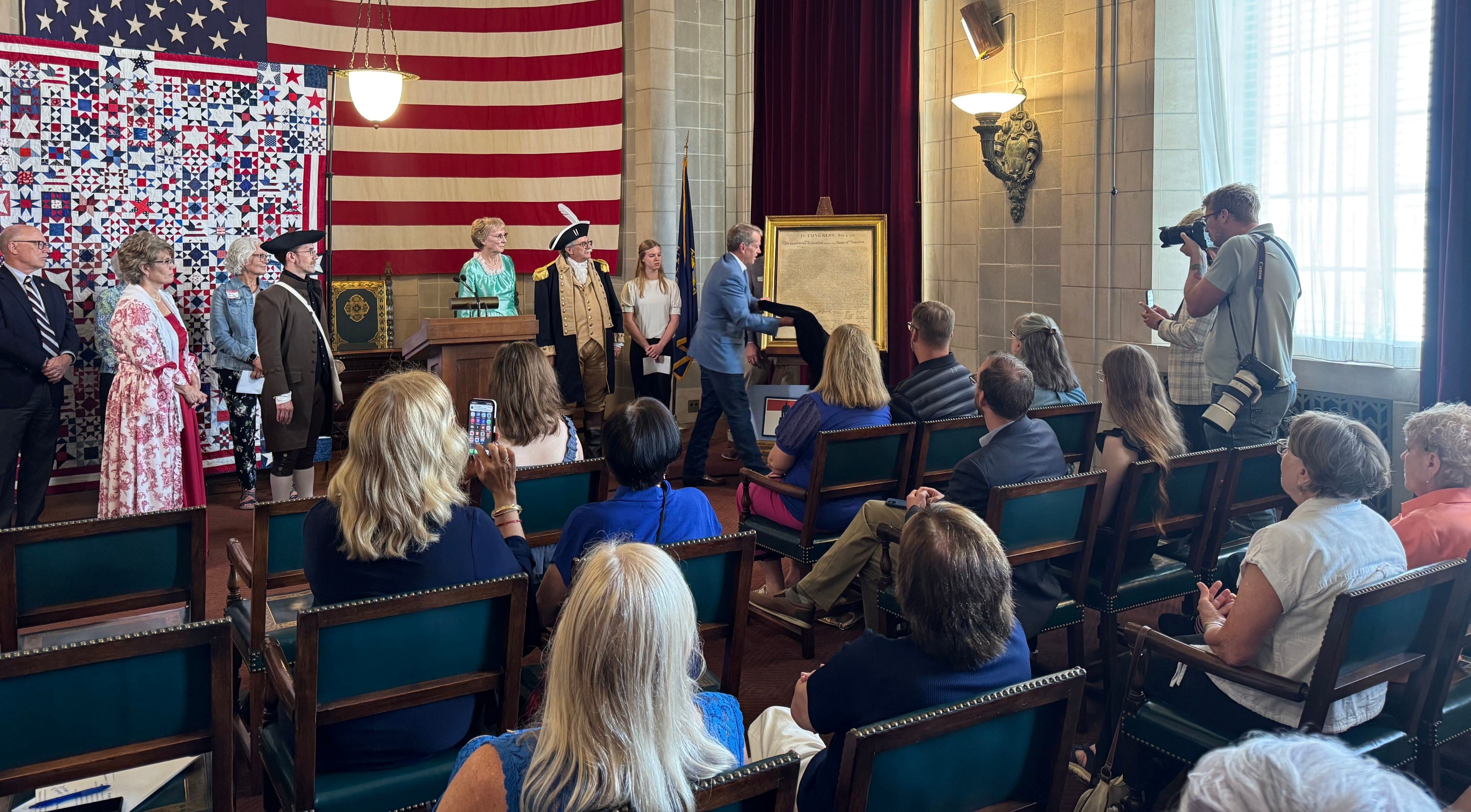 Governor Jim Pillen, flanked by members of the Sons and Daughters of the American Revolution, as well as others, unveils a framed copy of the Declaration of Independence in front of a gathered audience. The quilt, with 250-star blocks colored red, white, and blue and a huge American flag are in the background. 