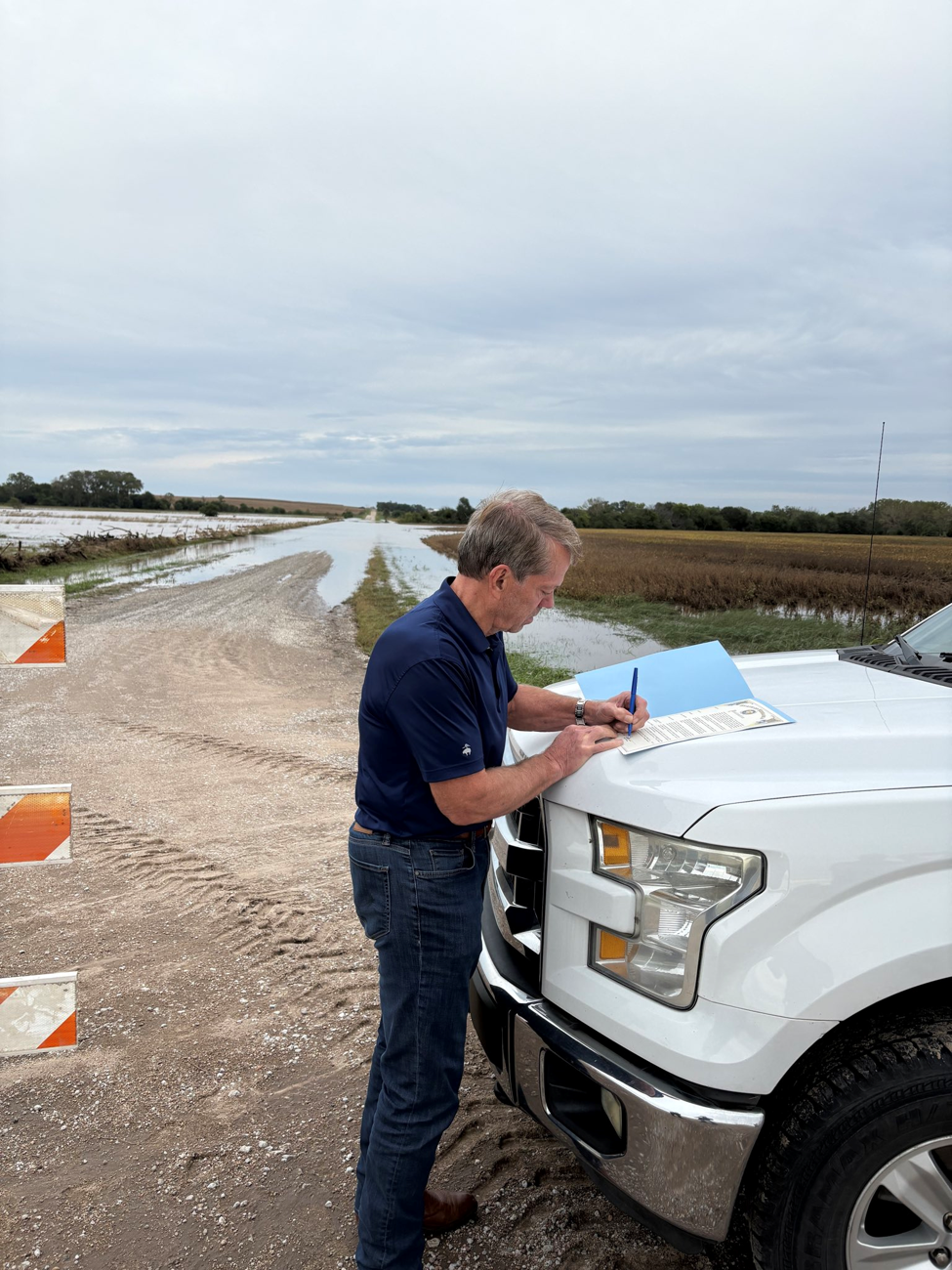 Gov. Pillen signs emergency proclamation on the hood of a white Emergency Management pickup truck while surveying a couple of water covered fields and gravel road in southeast Nebraska due to flooding.