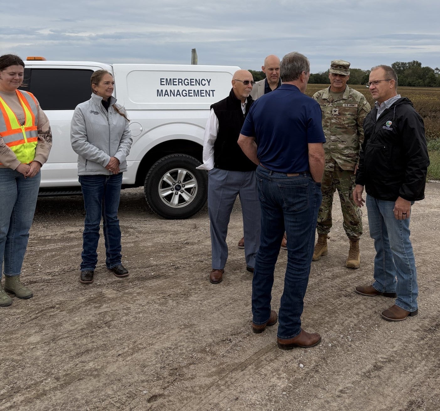 Governor briefed on flooding conditions by six Emergency Management personnel. A white Emergency Management pickup parked on a gravel road is in the background. 